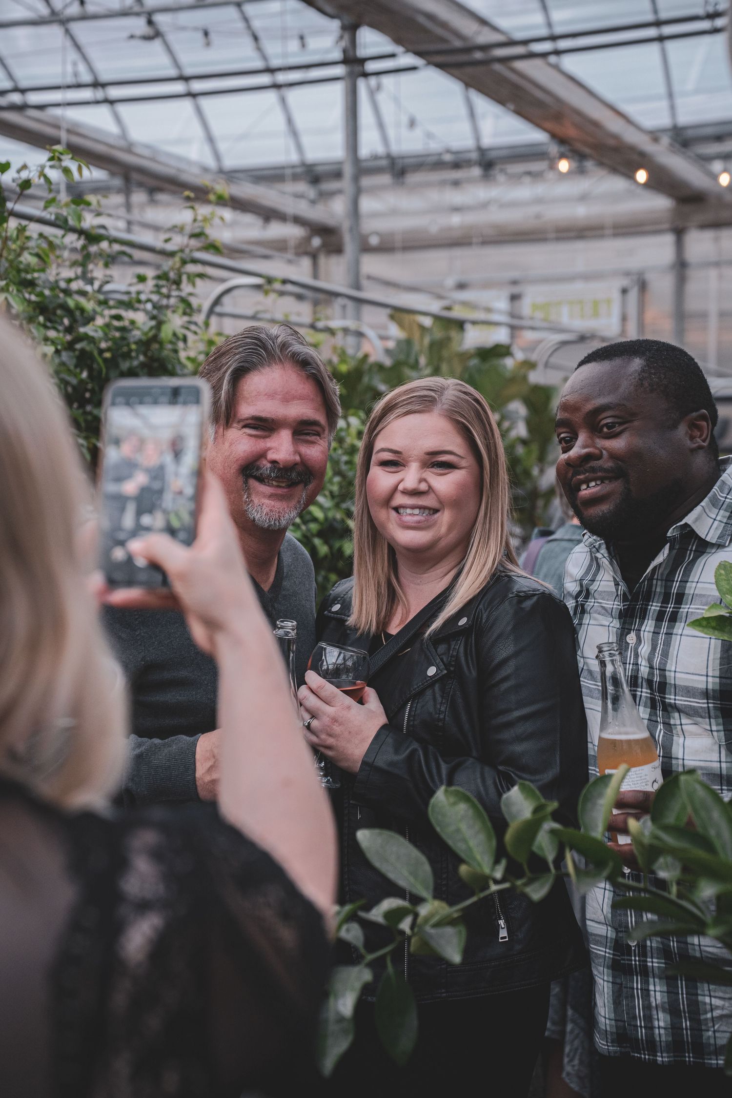 Three smiling people pose for a photo at a greenhouse social event while someone captures the moment on a smartphone.