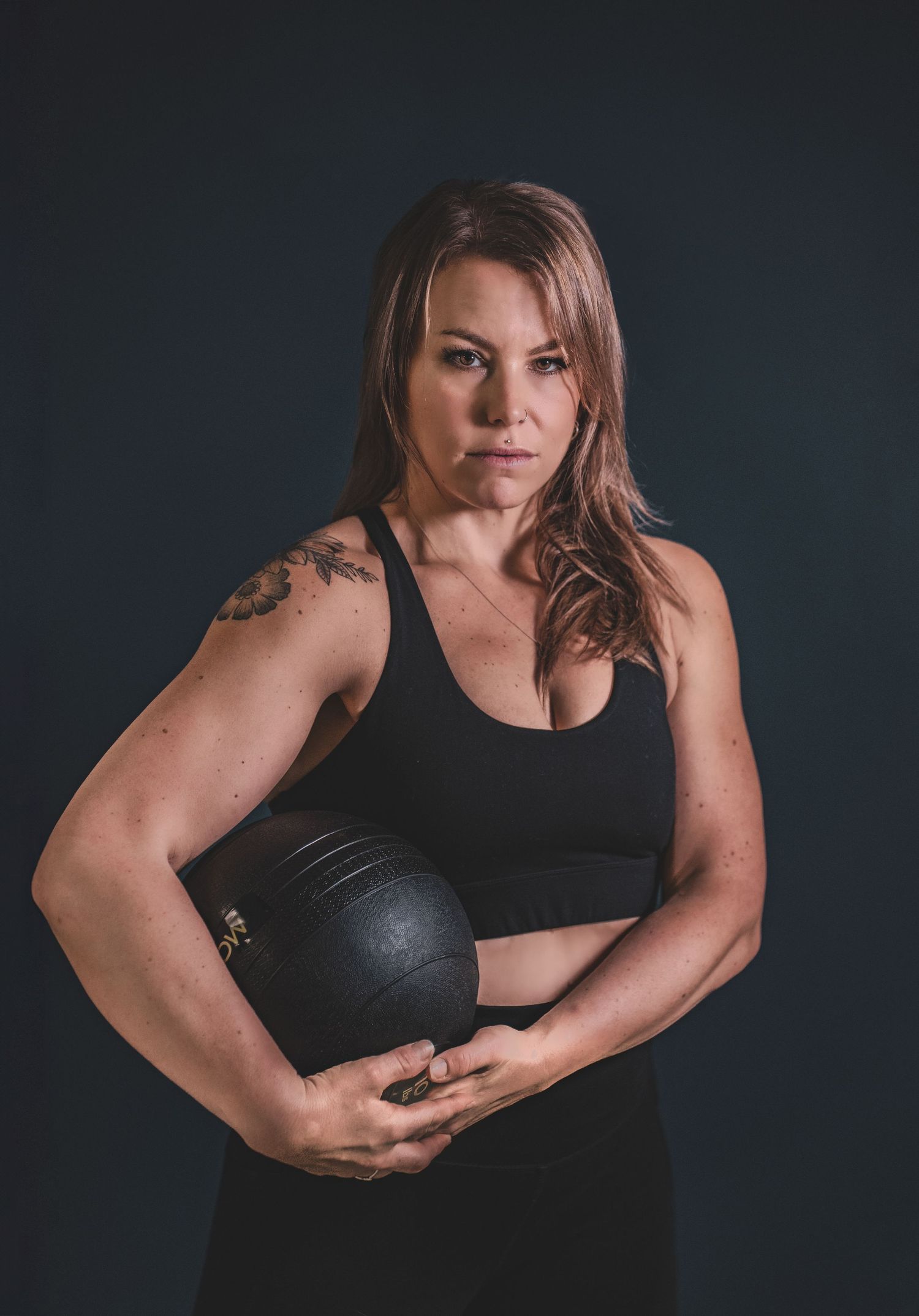 A muscular woman with a tattoo holds a medicine ball, posing confidently against a dark background during a branding photoshoot.