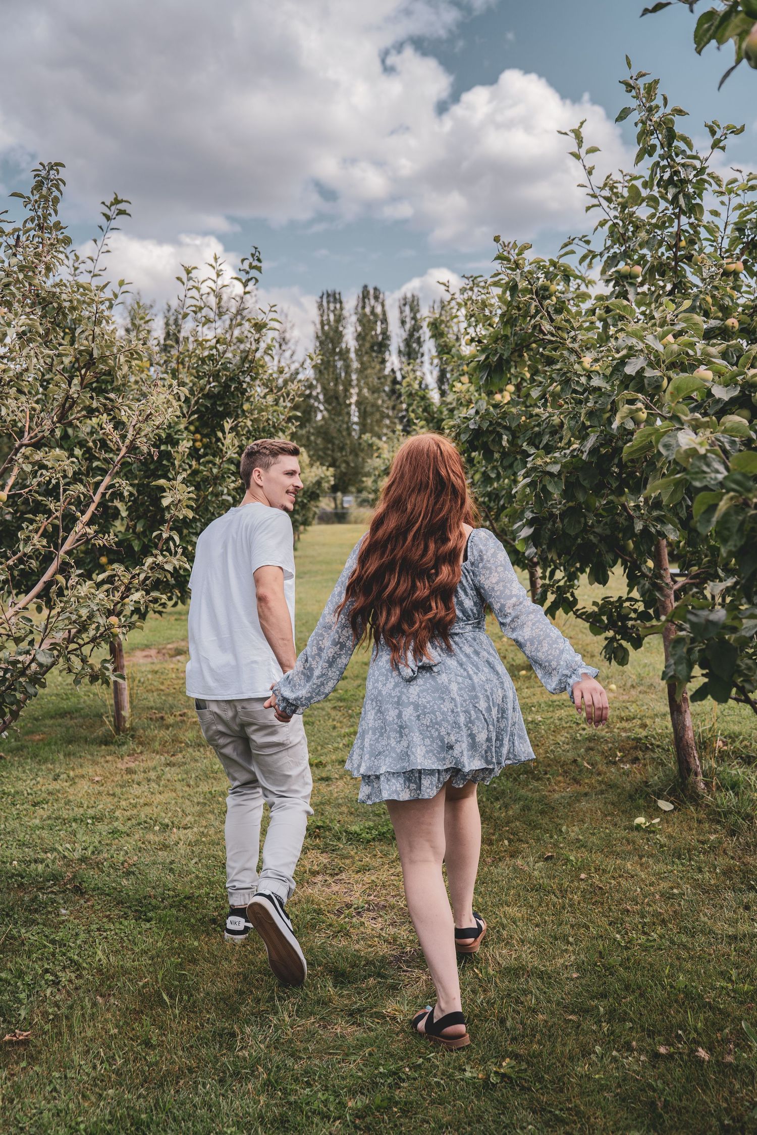 Couple walking hand in hand through a lush green apple orchard on a cloudy day.