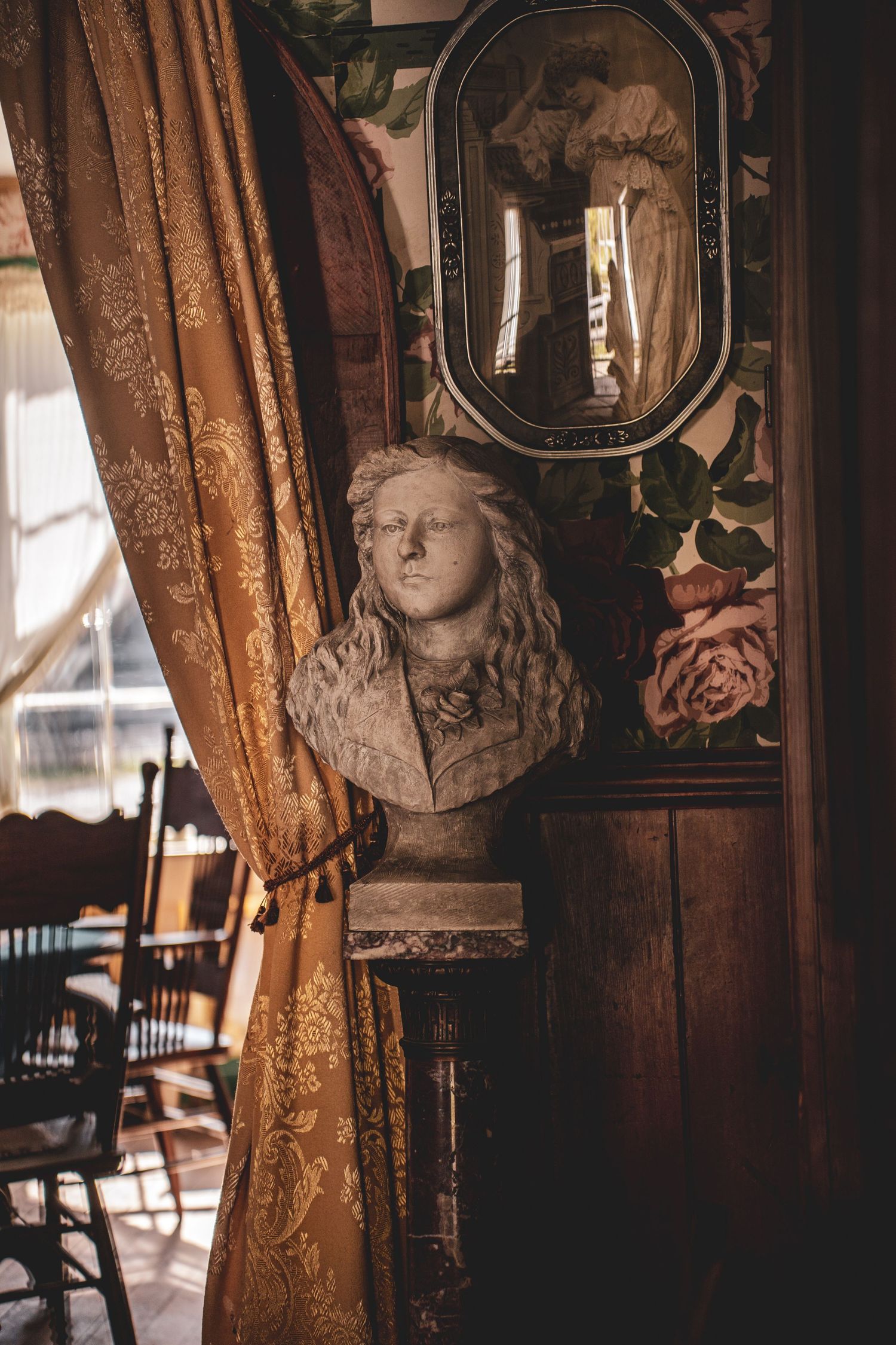 Antique bust sculpture displayed beside golden drapes and a ornate mirror in a vintage interior.