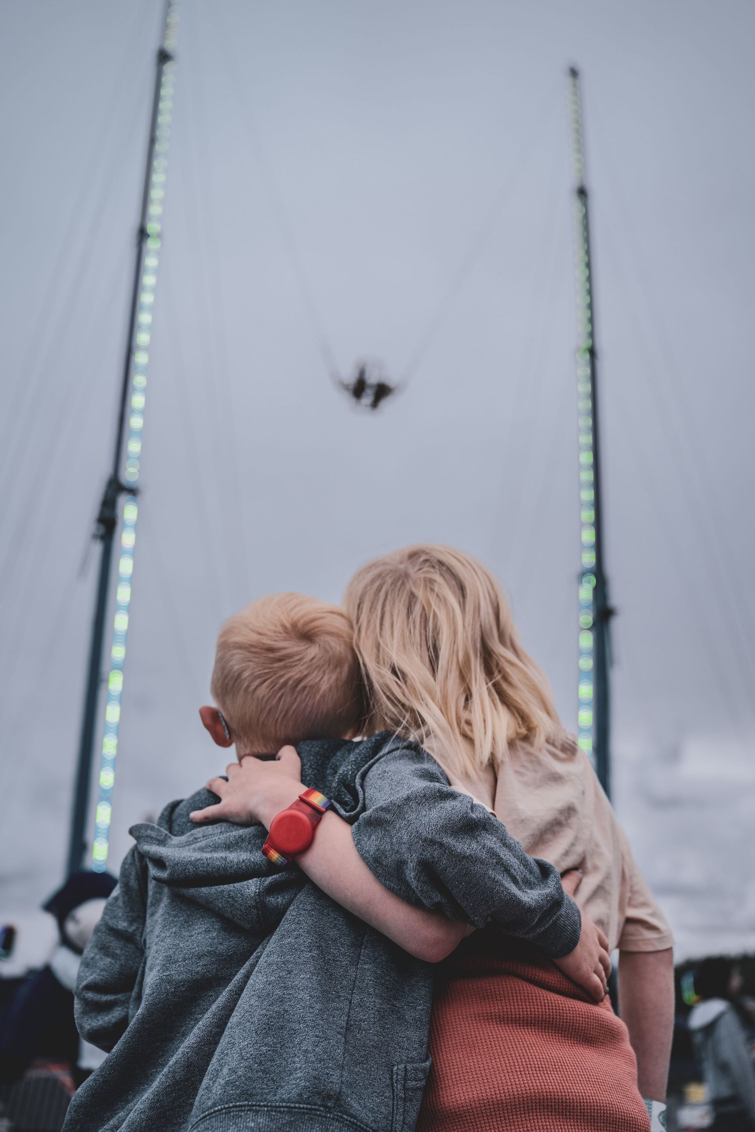 Two children hugging while watching a bungee jump attraction at an outdoor fairground event.