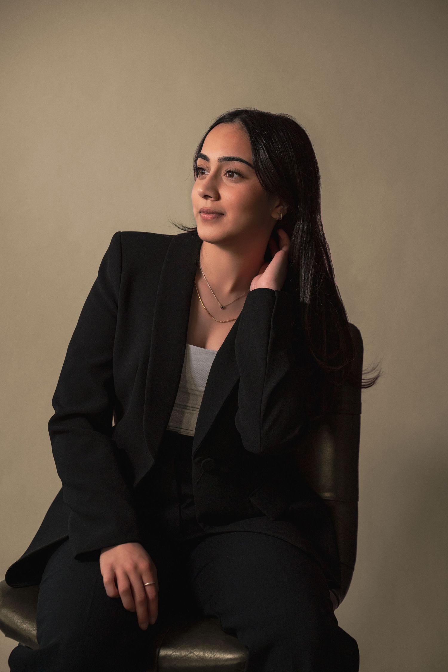 Young woman in black suit touching hair while seated against beige studio backdrop during her real estate headshot photoshoot.