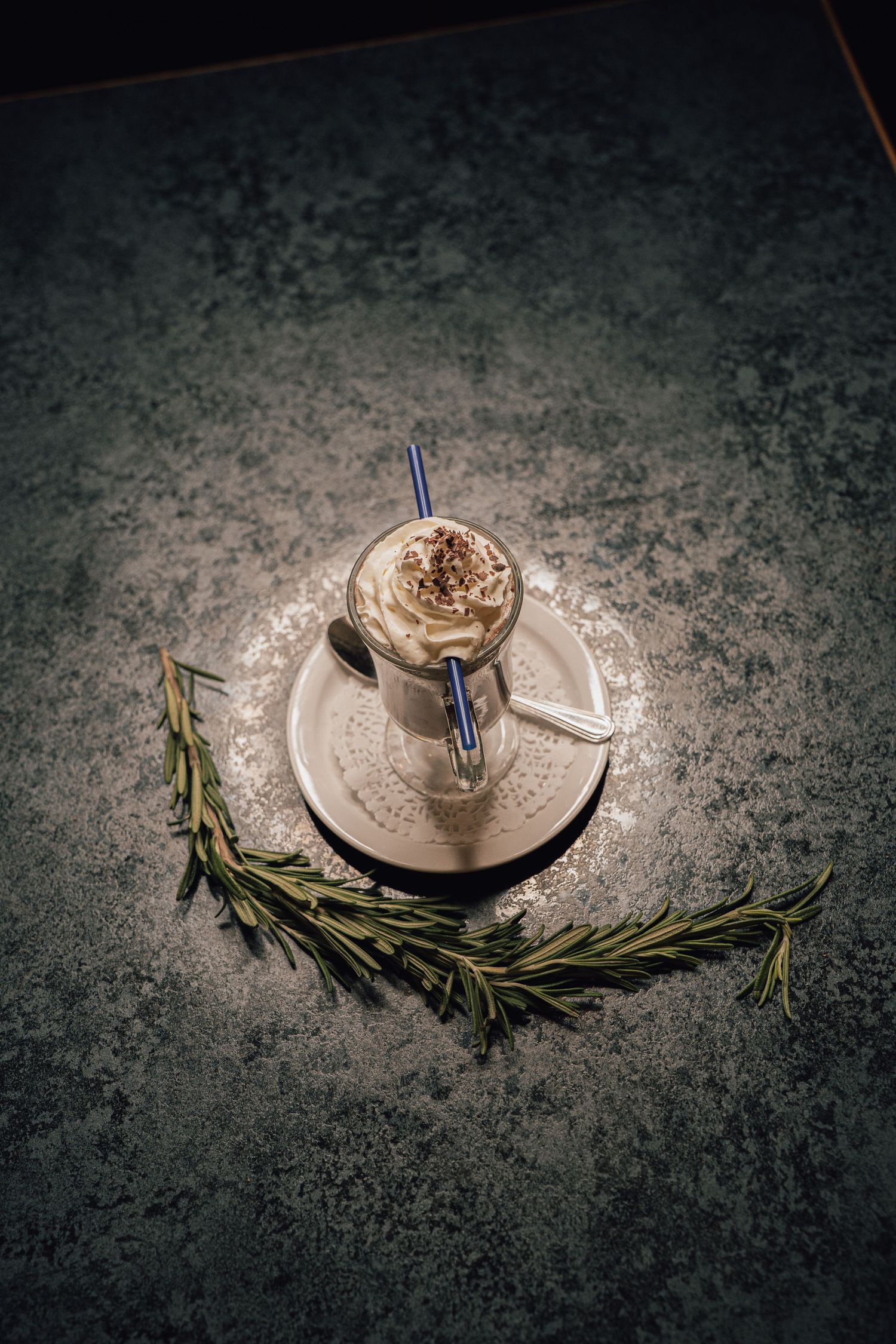 Top-down view of a whipped cream coffee drink on a saucer with rosemary sprigs on dark surface.