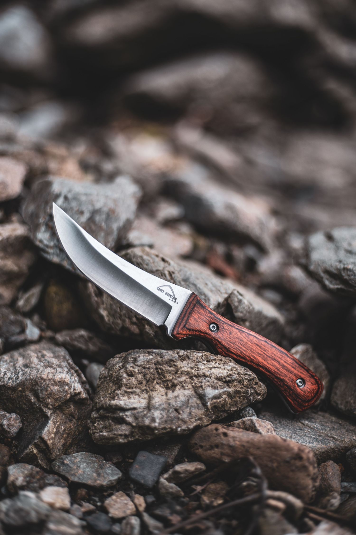 Product photography of Fixed blade hunting knife with wood handle resting on rugged rocky terrain outdoors.