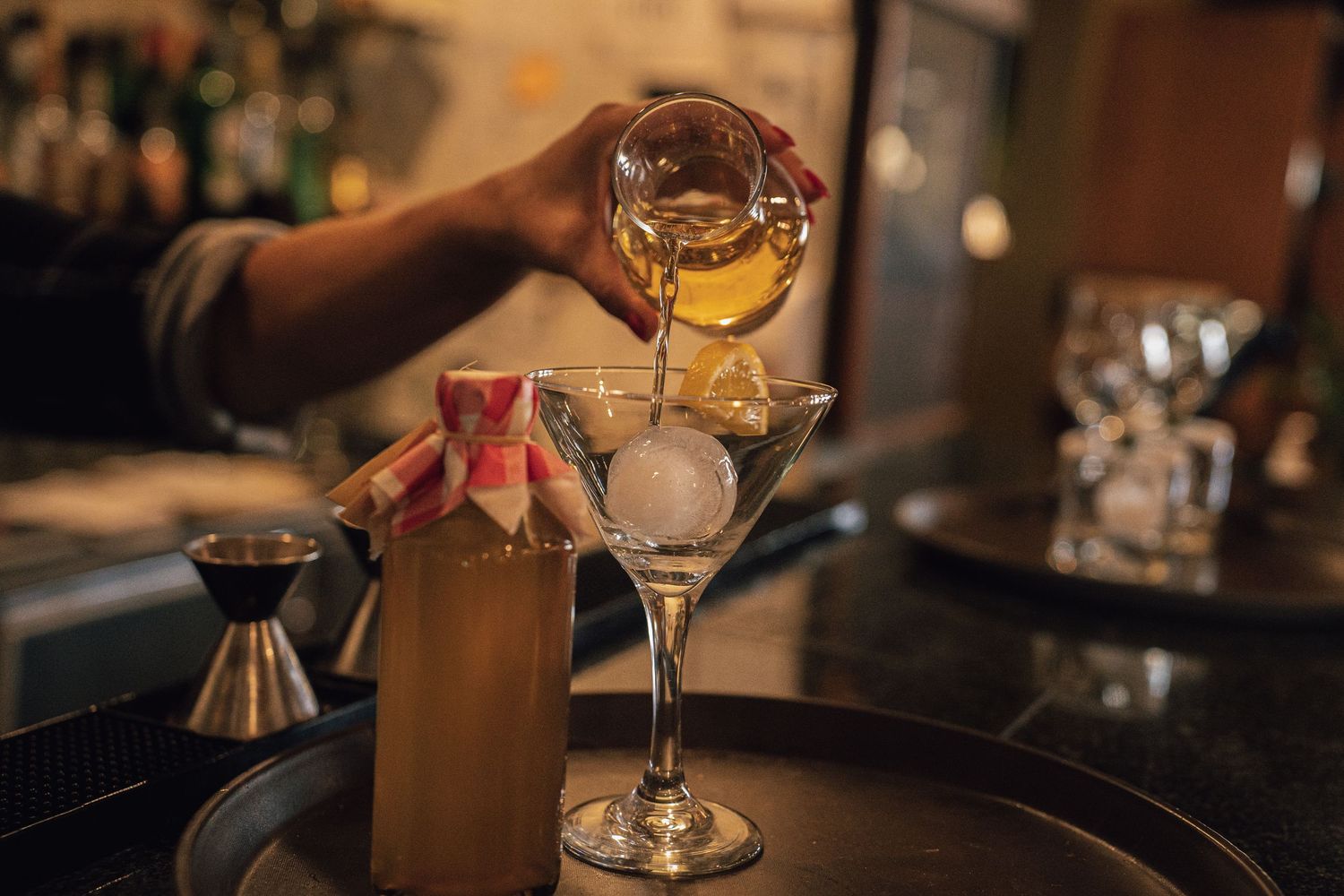 Bartender pouring amber cocktail into a chilled martini glass with a large ice sphere.