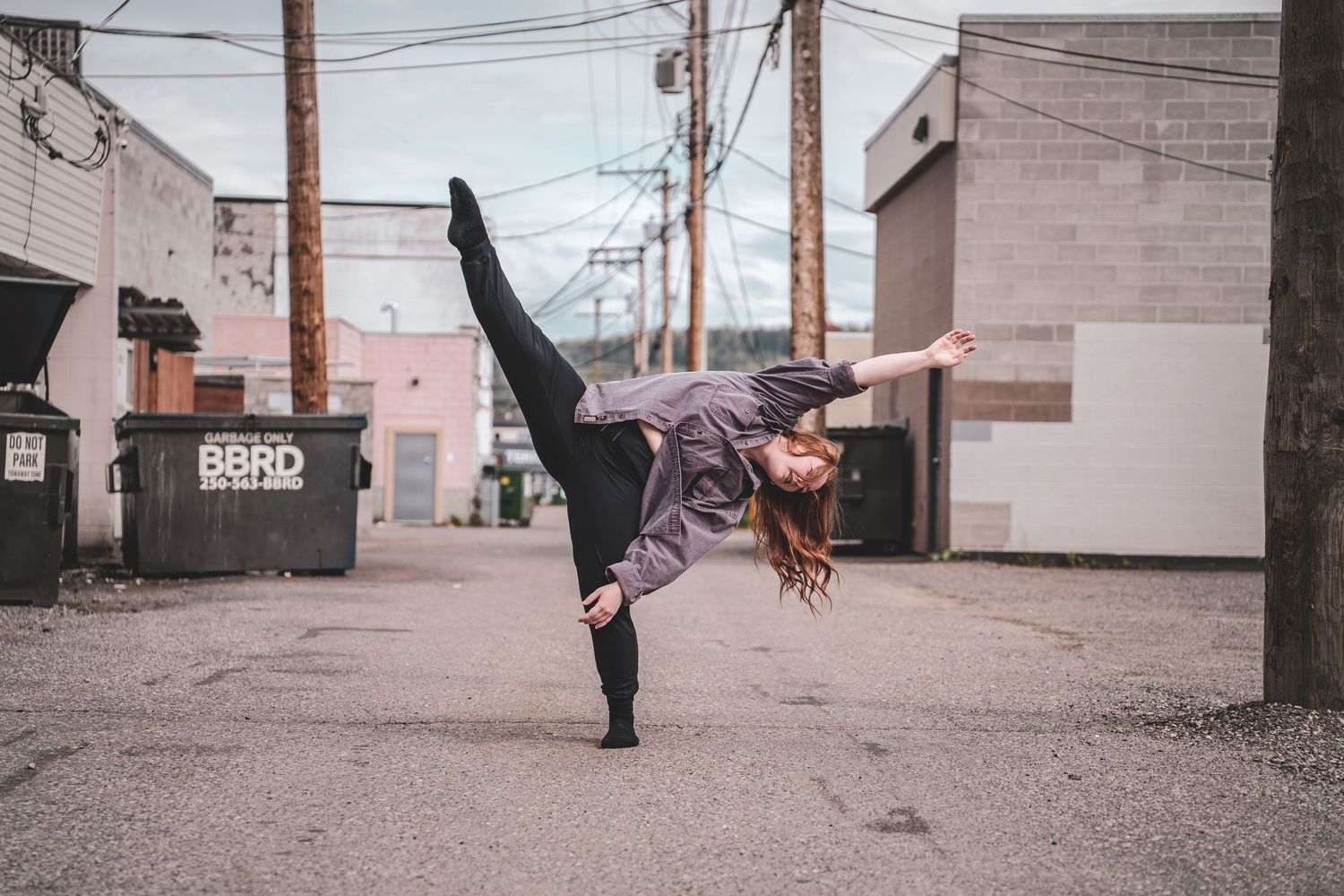 A female dancer performs a high kick balance pose outdoors in an urban alley setting during branding session.