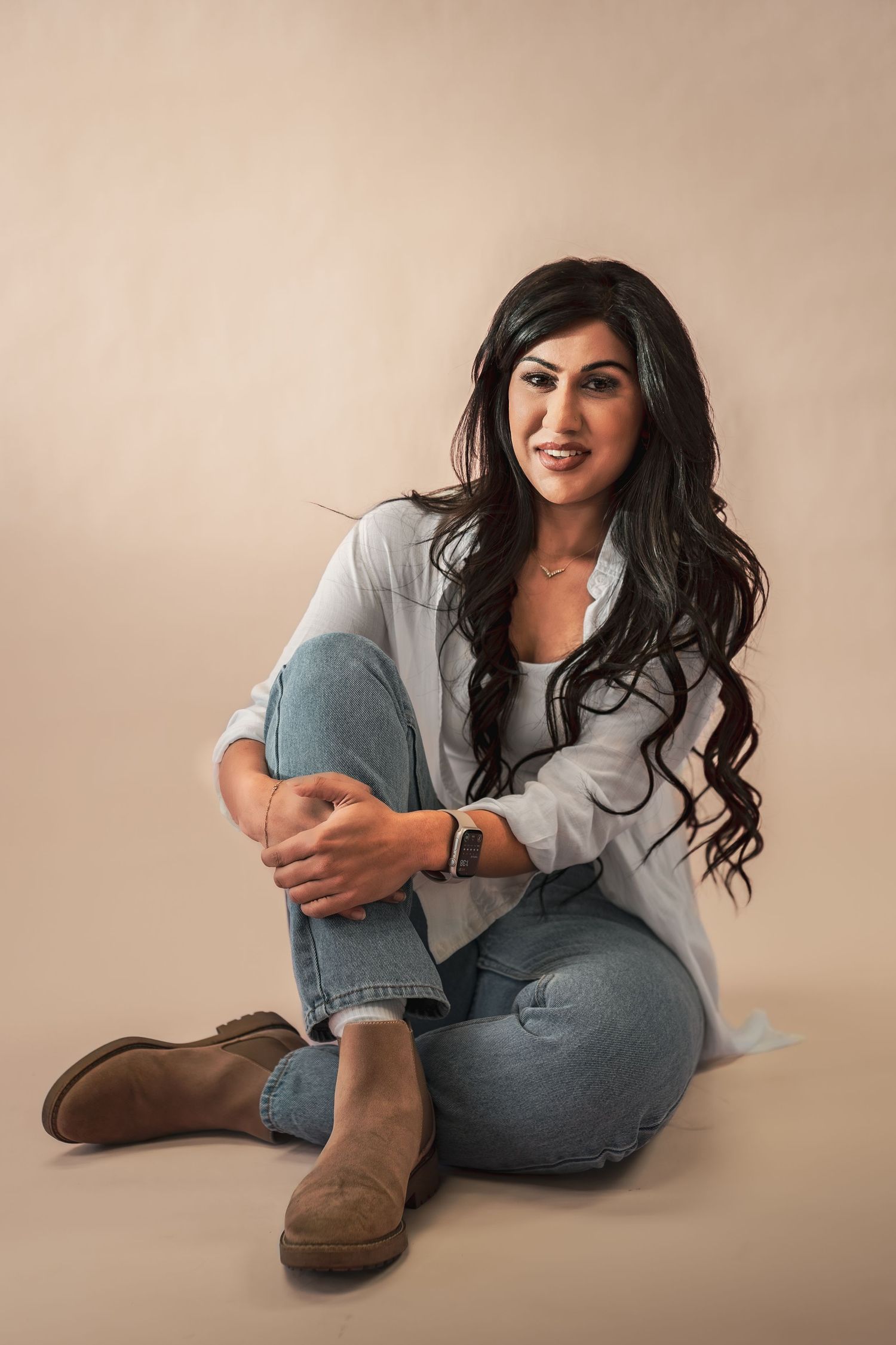 A smiling woman with long dark wavy hair sits on the floor in jeans and a white shirt on a beige backdrop during her business branding photo