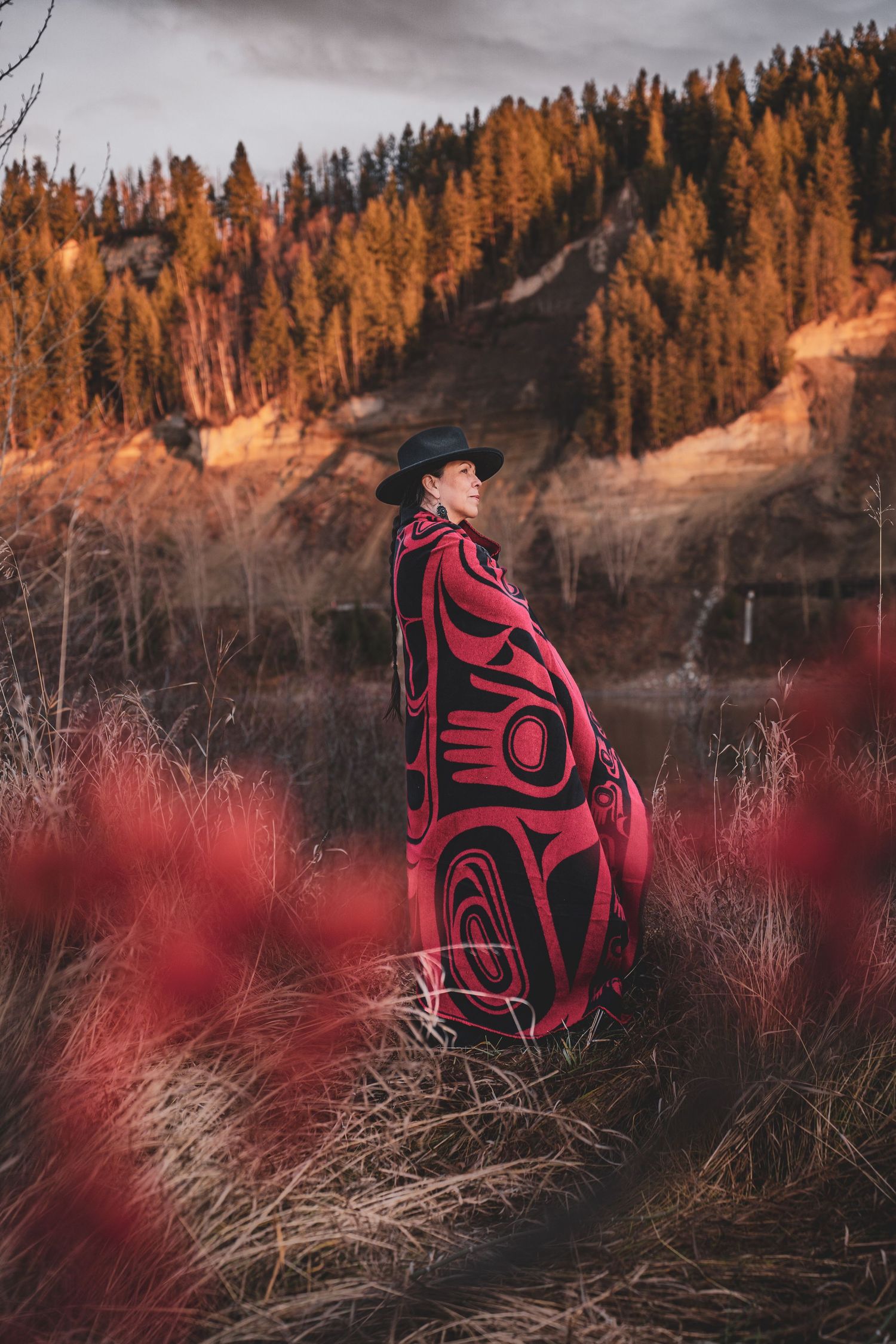 Kym Gouchie wrapped in red Indigenous patterned blanket wearing black hat, standing in autumn field by river creative photoshoot.