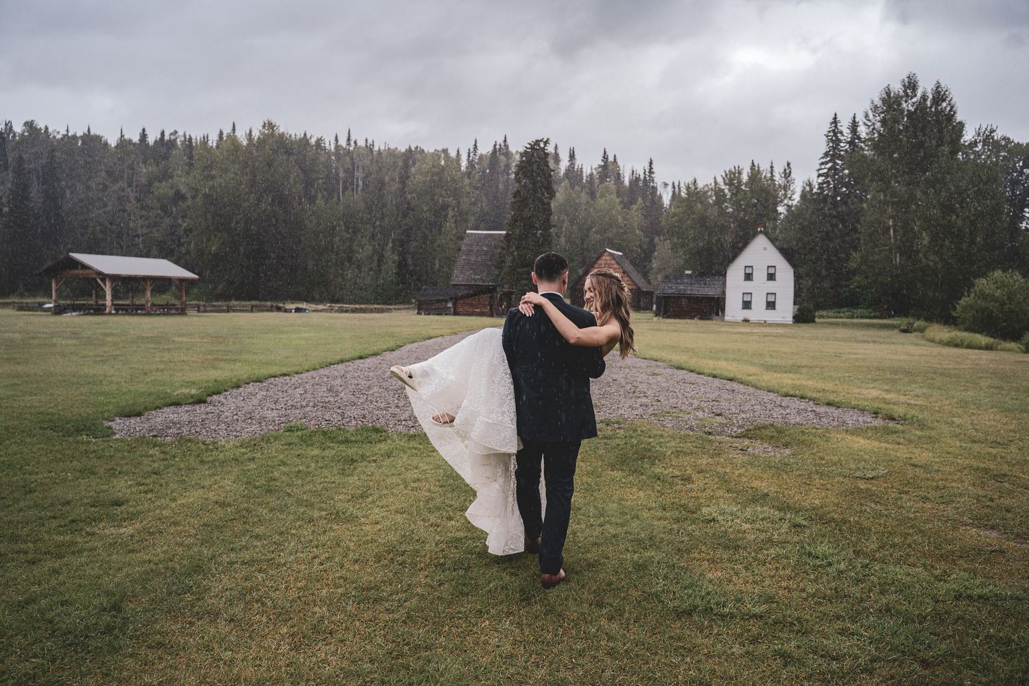 Groom lifting bride in a green field with rustic buildings and forest in the background.