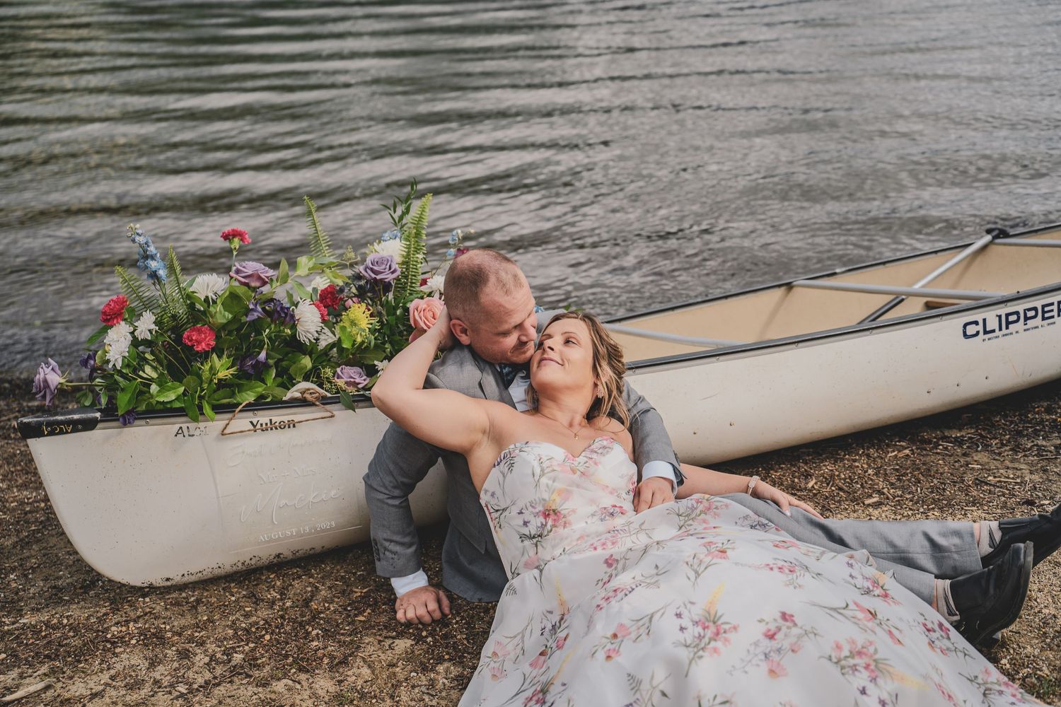 Couple relaxing by a canoe filled with colorful flowers on a lakeside shore after their dock elopement offered by photography.