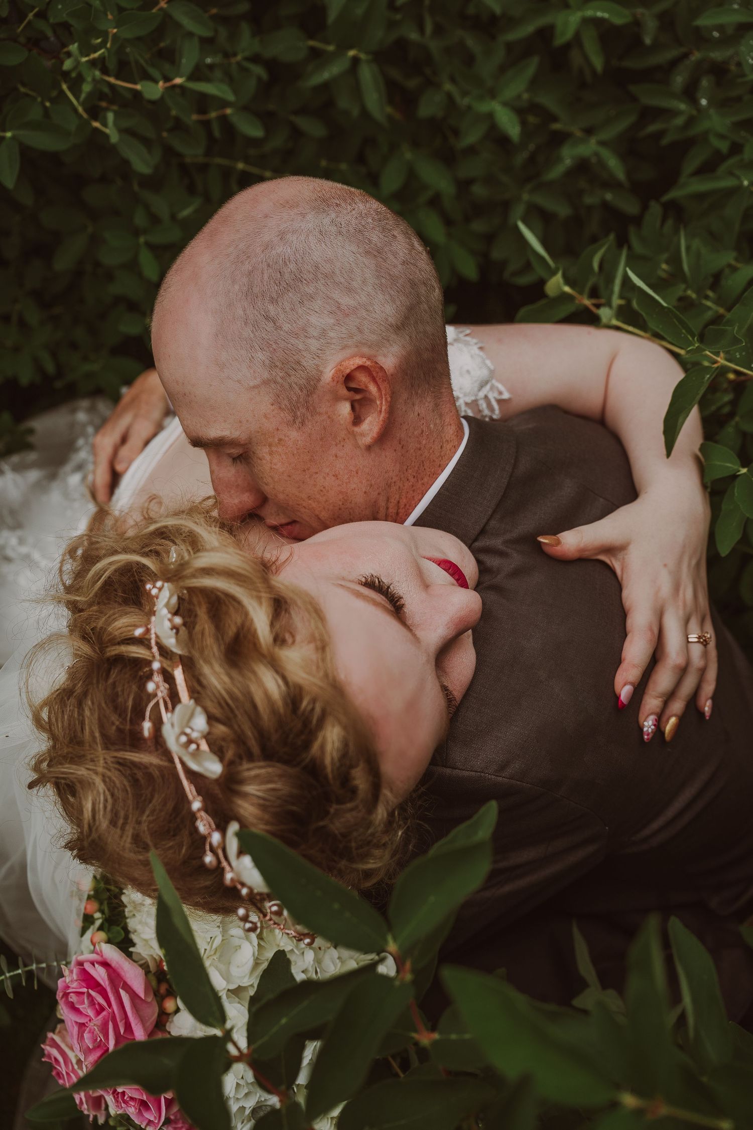 Bride and groom sharing intimate embrace surrounded by lush green foliage during outdoor wedding at Northern Lights Winery, Prince George, B