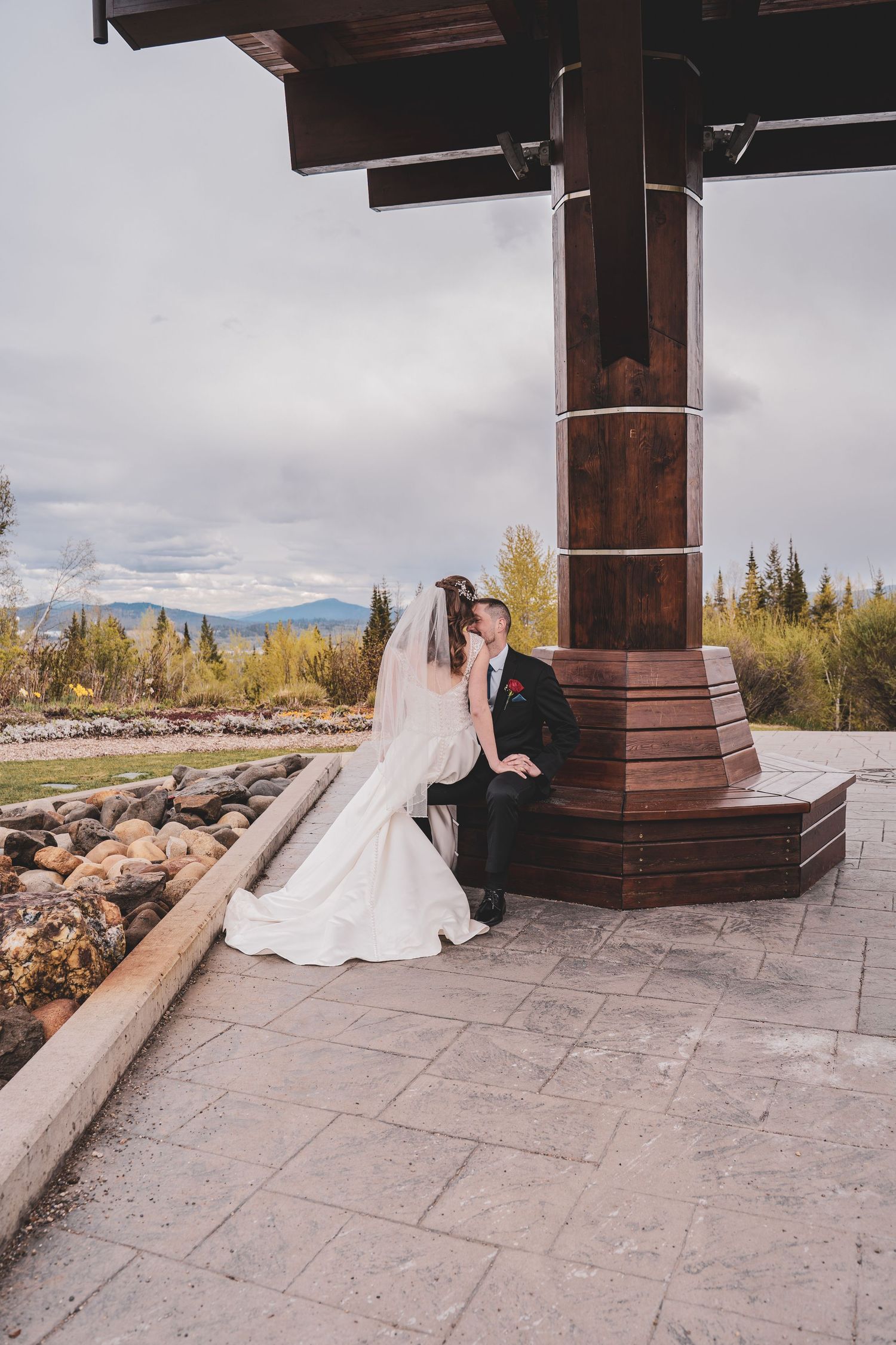 Bride and groom share a romantic kiss seated beside a large wooden pavilion post with mountain views.