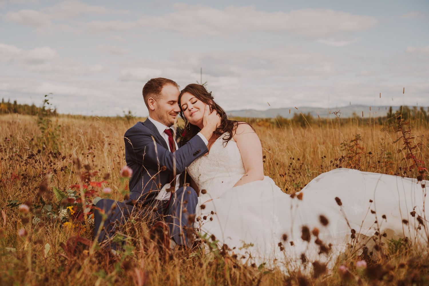 Bride and groom sitting in golden field, groom tenderly cradling bride's face with mountains in background.