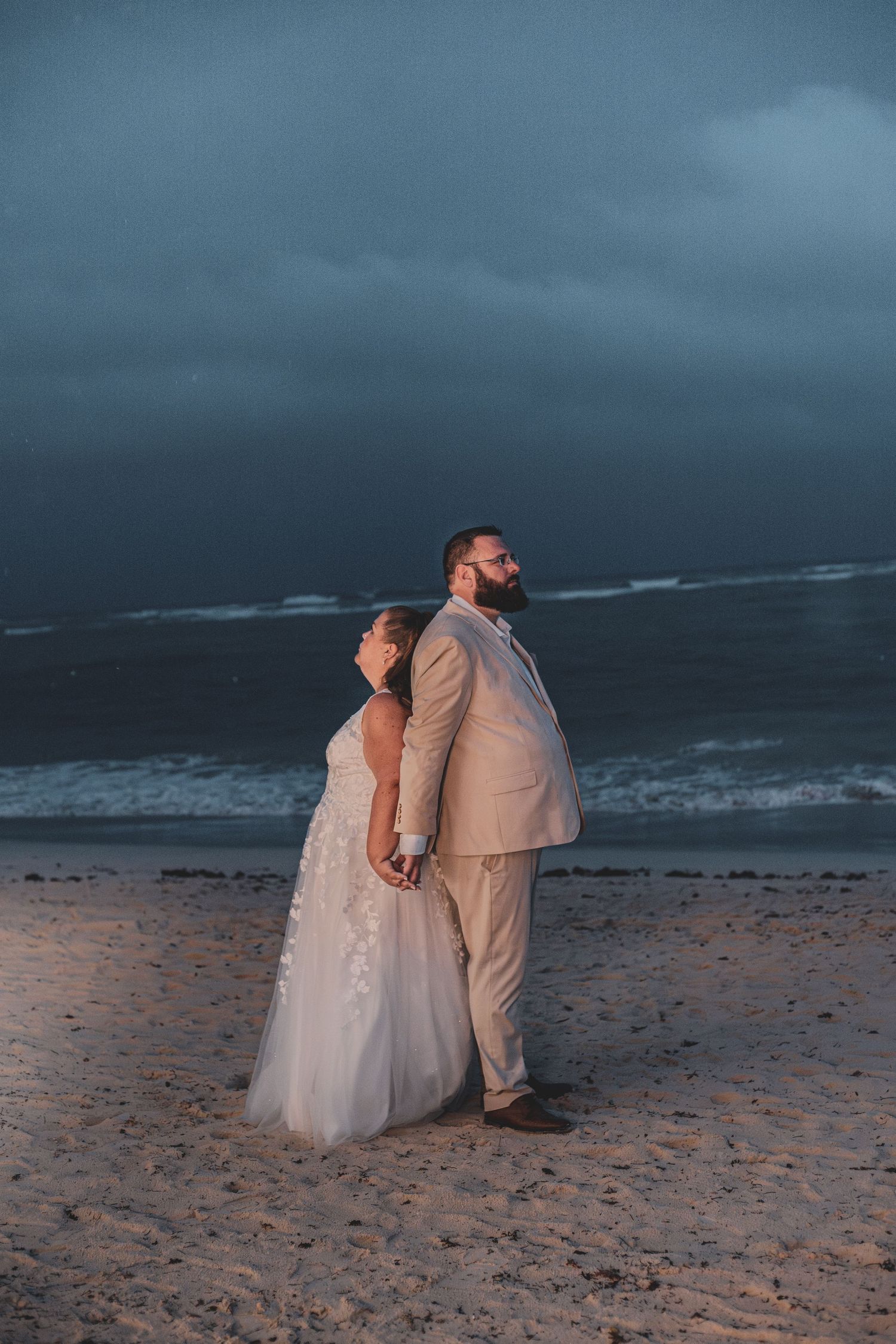 Bride and groom standing back-to-back on a sandy beach under dramatic stormy skies.