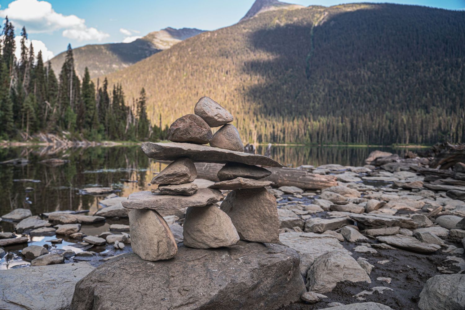 Stacked rock cairn balancing beside a serene mountain lake surrounded by pine trees and peaks.
