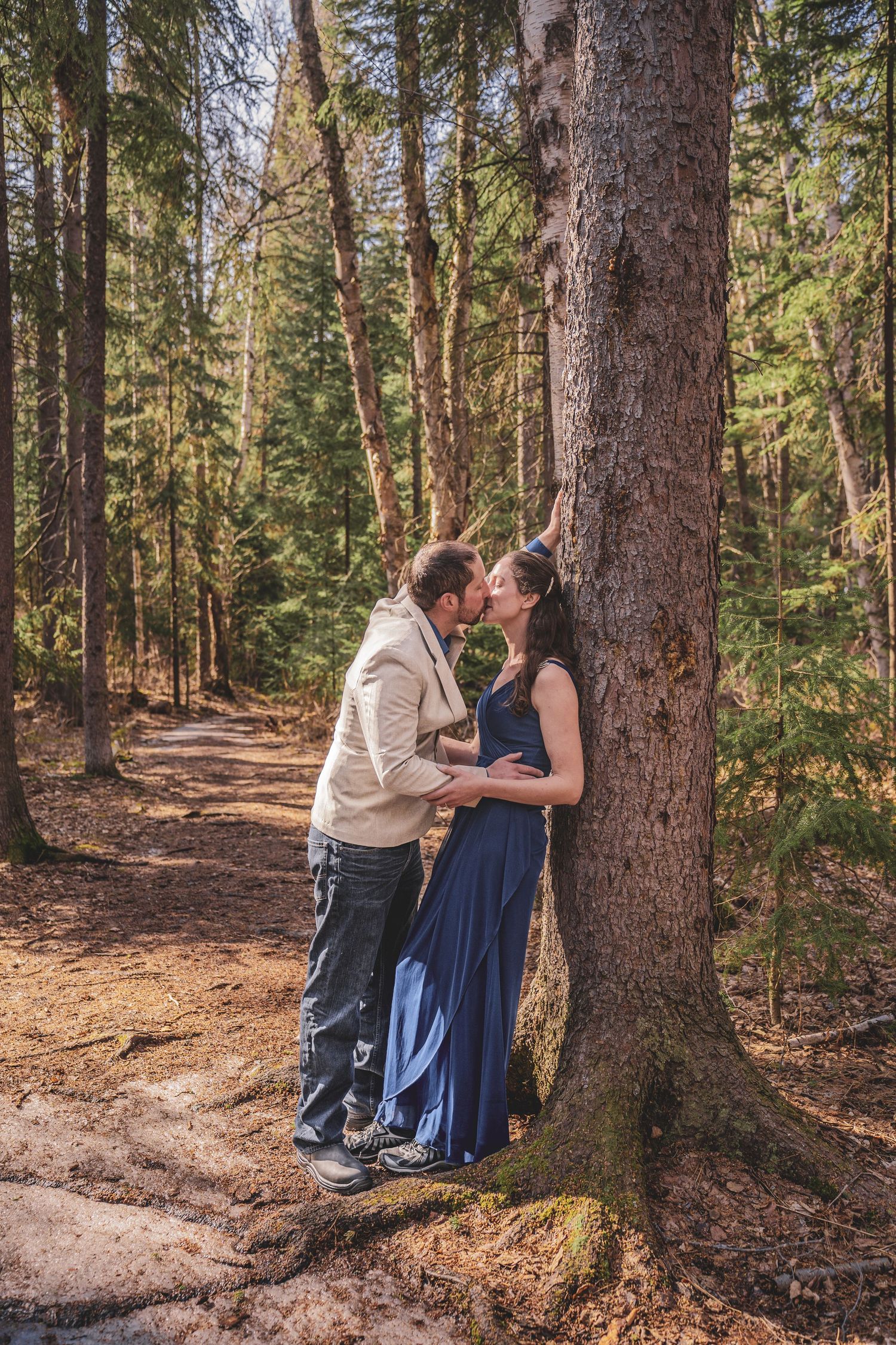Couple sharing a romantic kiss leaning against tall tree trunk in sunny pine forest.