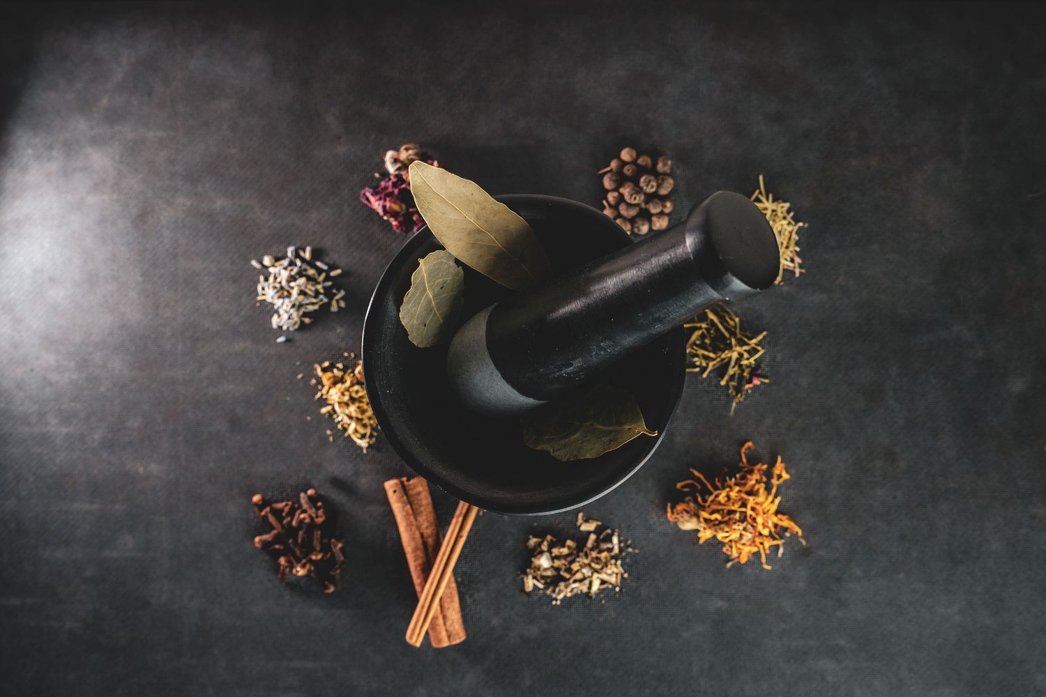 Product photography of Black mortar and pestle surrounded by assorted dried herbs and spices on dark surface from shop in PG.