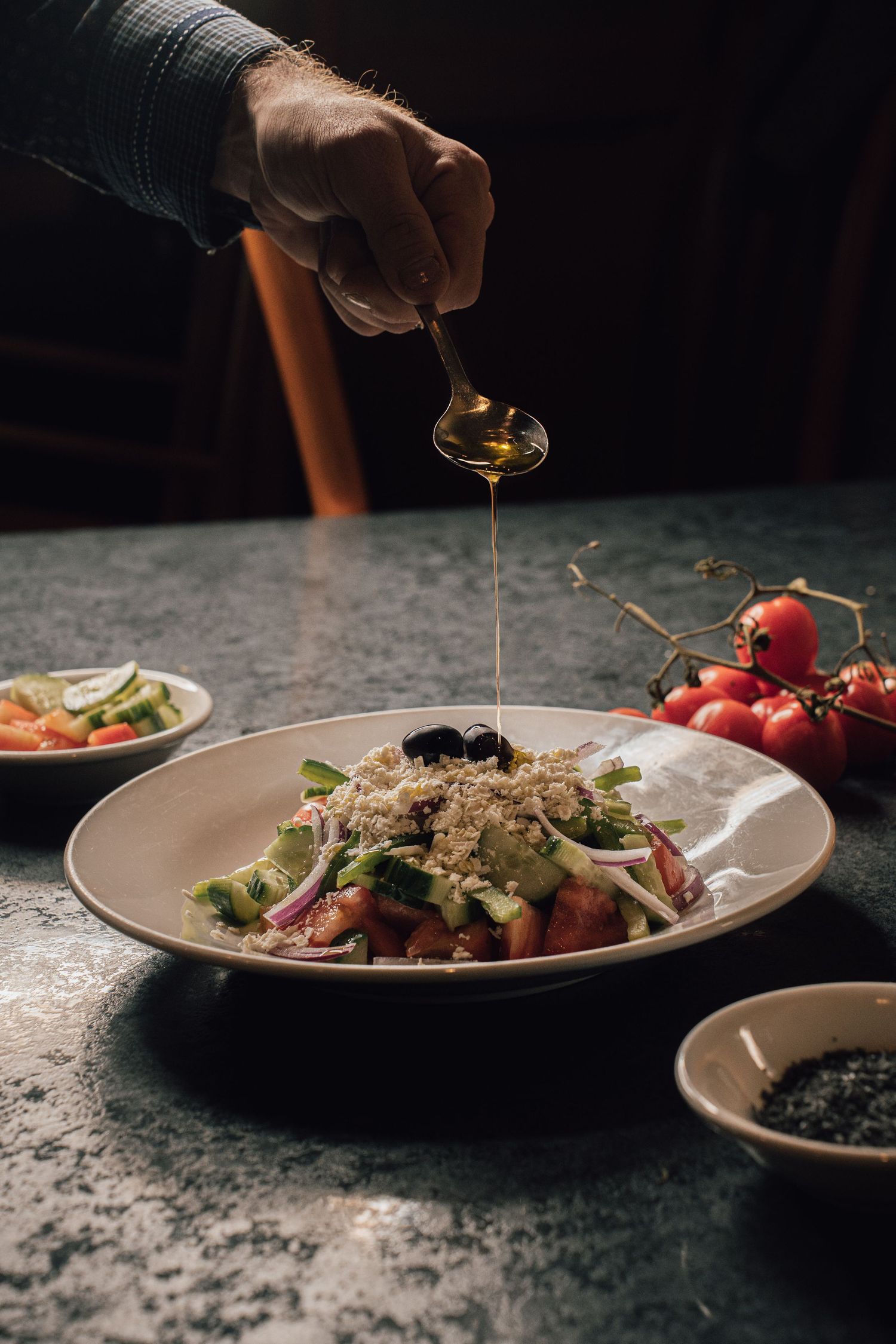 Chef drizzling olive oil over a fresh quinoa salad with vegetables and olives in a white bowl.