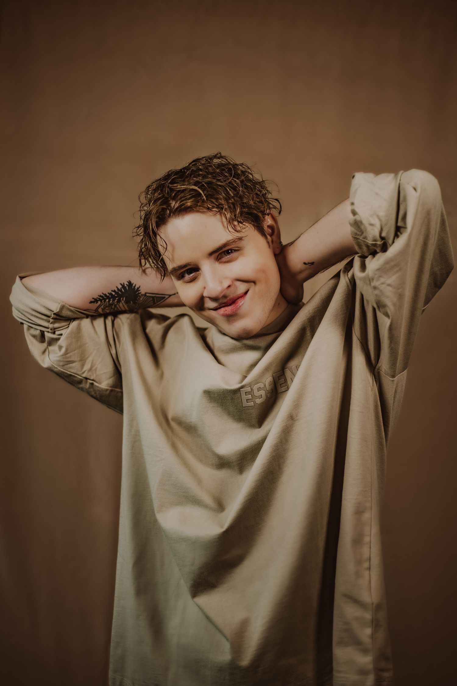A smiling young man with curly hair poses casually in an oversized beige shirt against a warm brown background during a modeling photoshoot