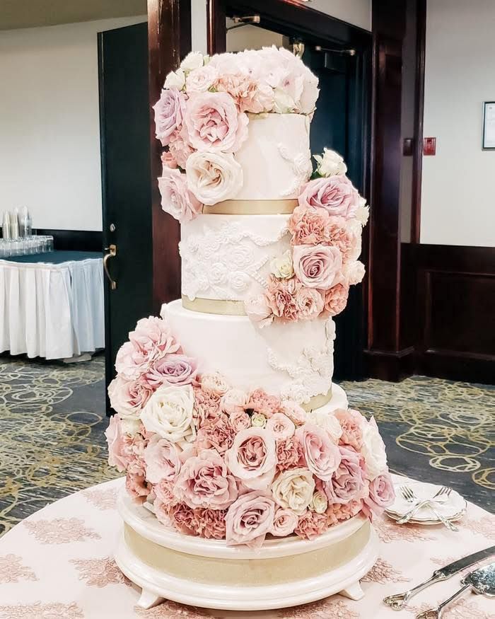 Four-tier white wedding cake adorned with lush pink and mauve roses with gold accent bands, offered by Prince george cake maker