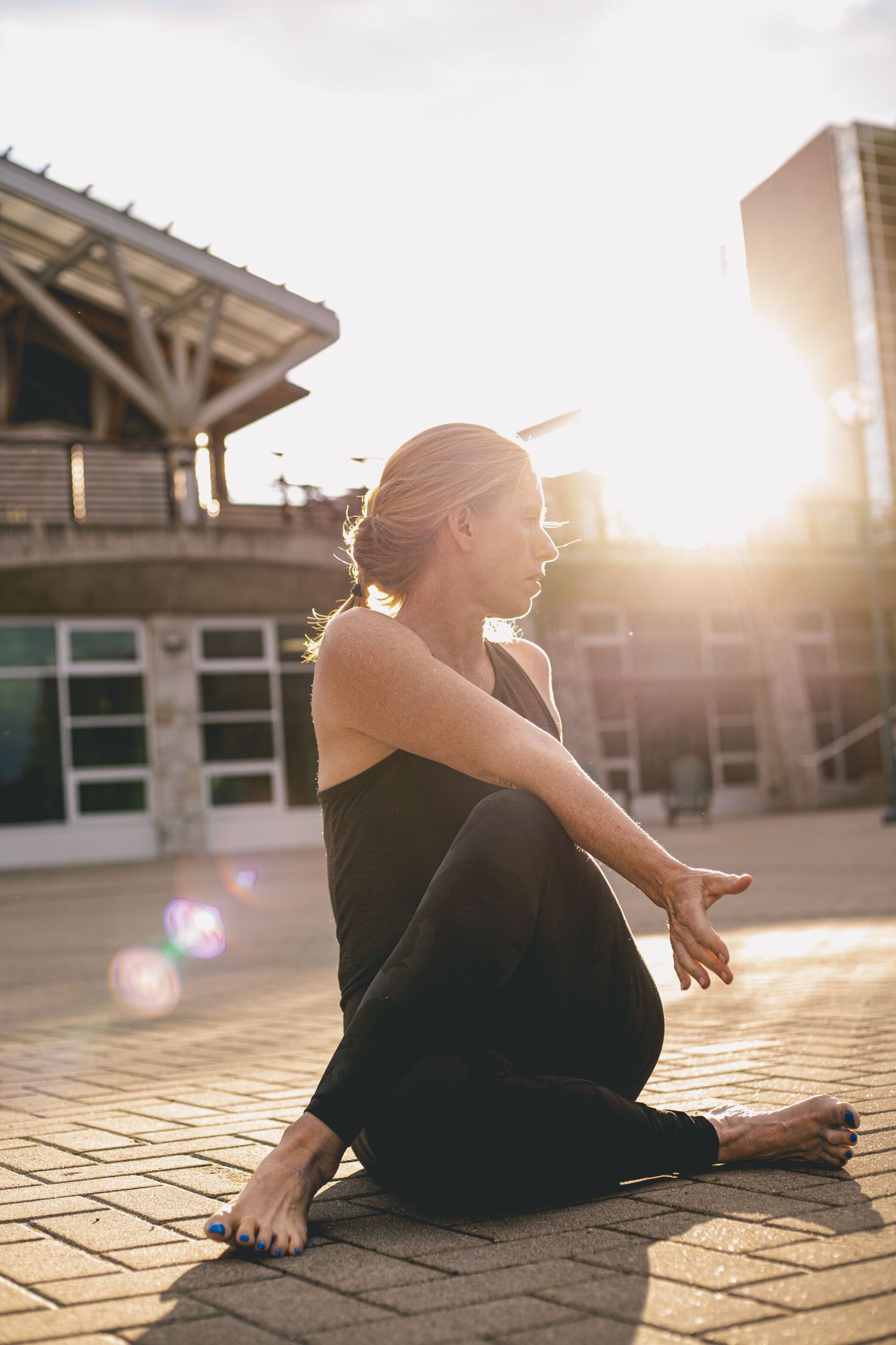 A barefoot woman in all-black activewear sits cross-legged on pavement in bright golden sunlight for her business photoshoot portfolio.