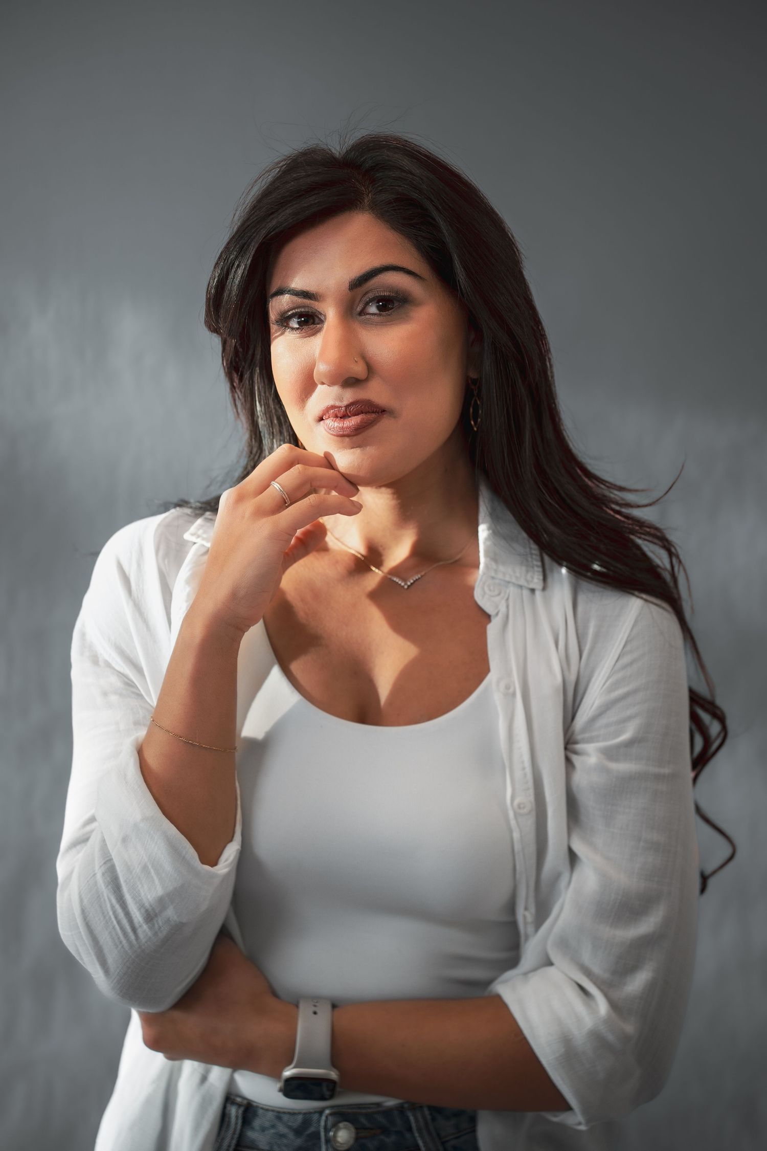 A confident woman with long dark hair poses in a white shirt and tank top against a gray background during her headshot photoshoot.