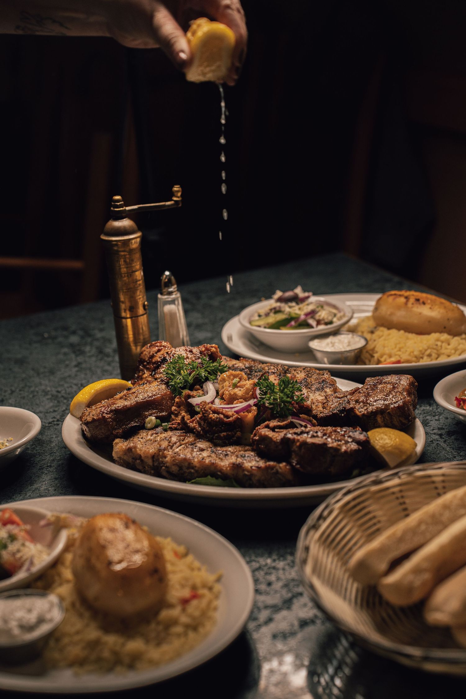 Hand squeezing lemon over a large platter of grilled mixed meats with sides and bread.