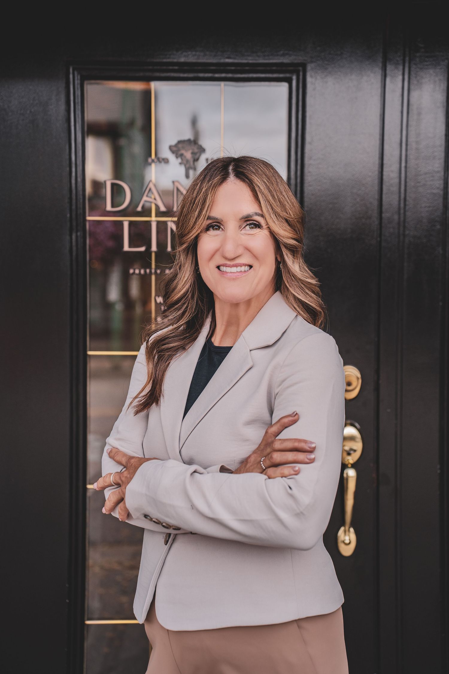 Professional woman in gray blazer smiling with arms crossed outside a boutique door.