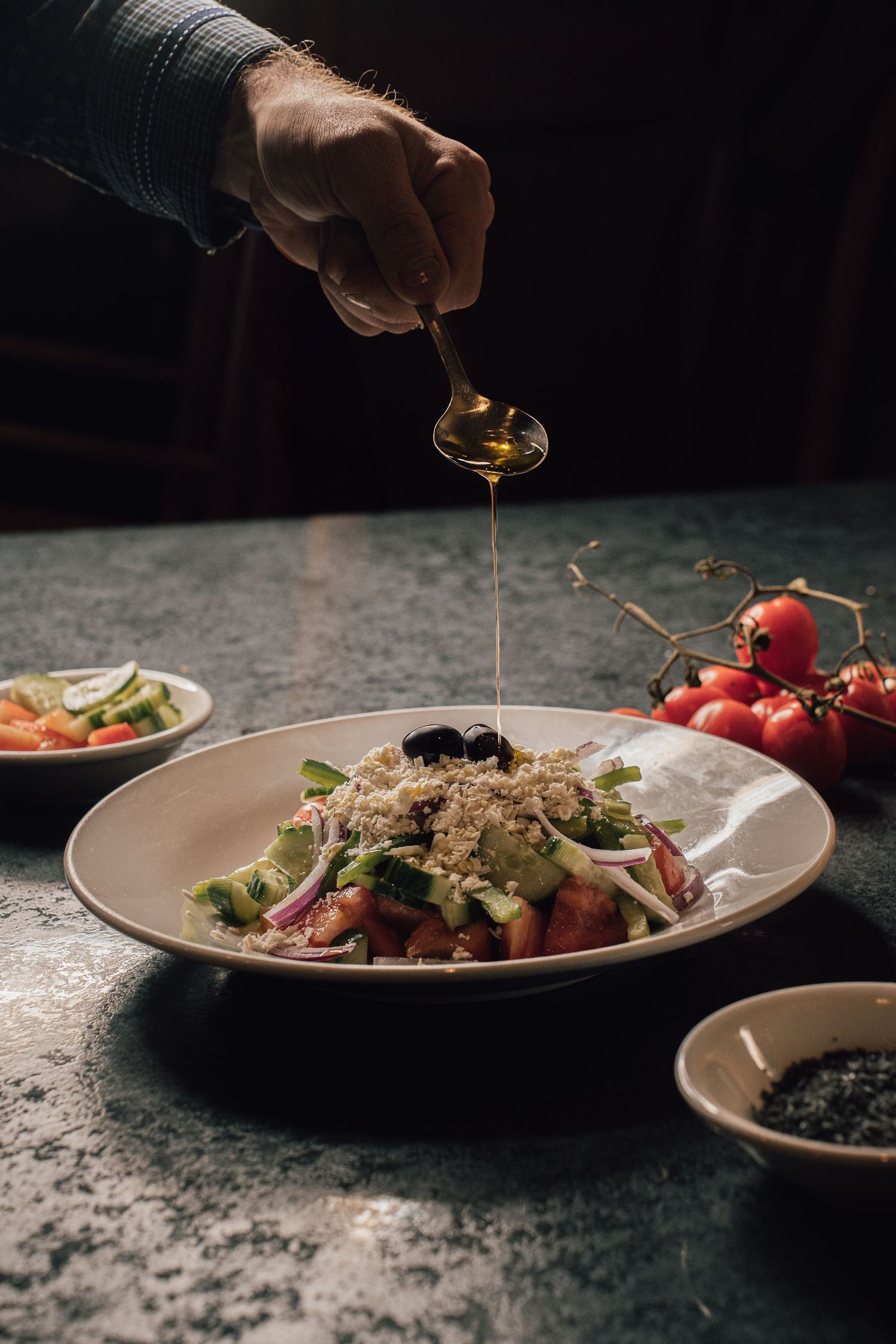Hand drizzling olive oil from spoon onto fresh Greek salad on dark background.