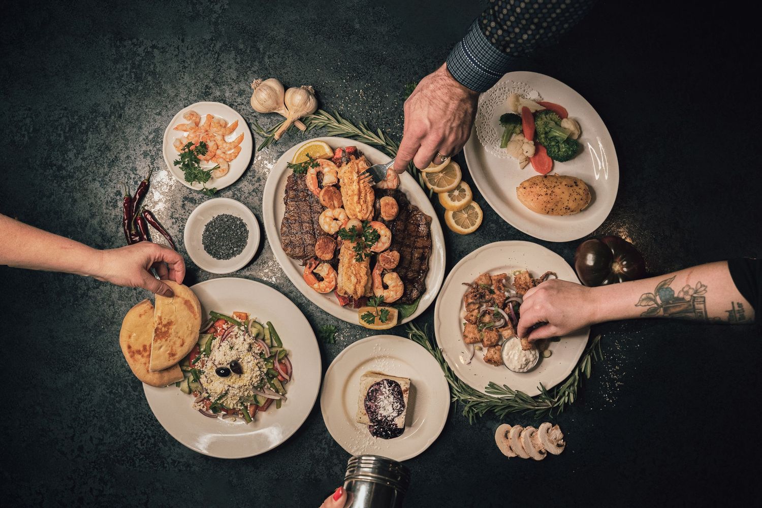 People reaching for dishes in a shared Mediterranean seafood feast with grilled meats and fresh salads.
