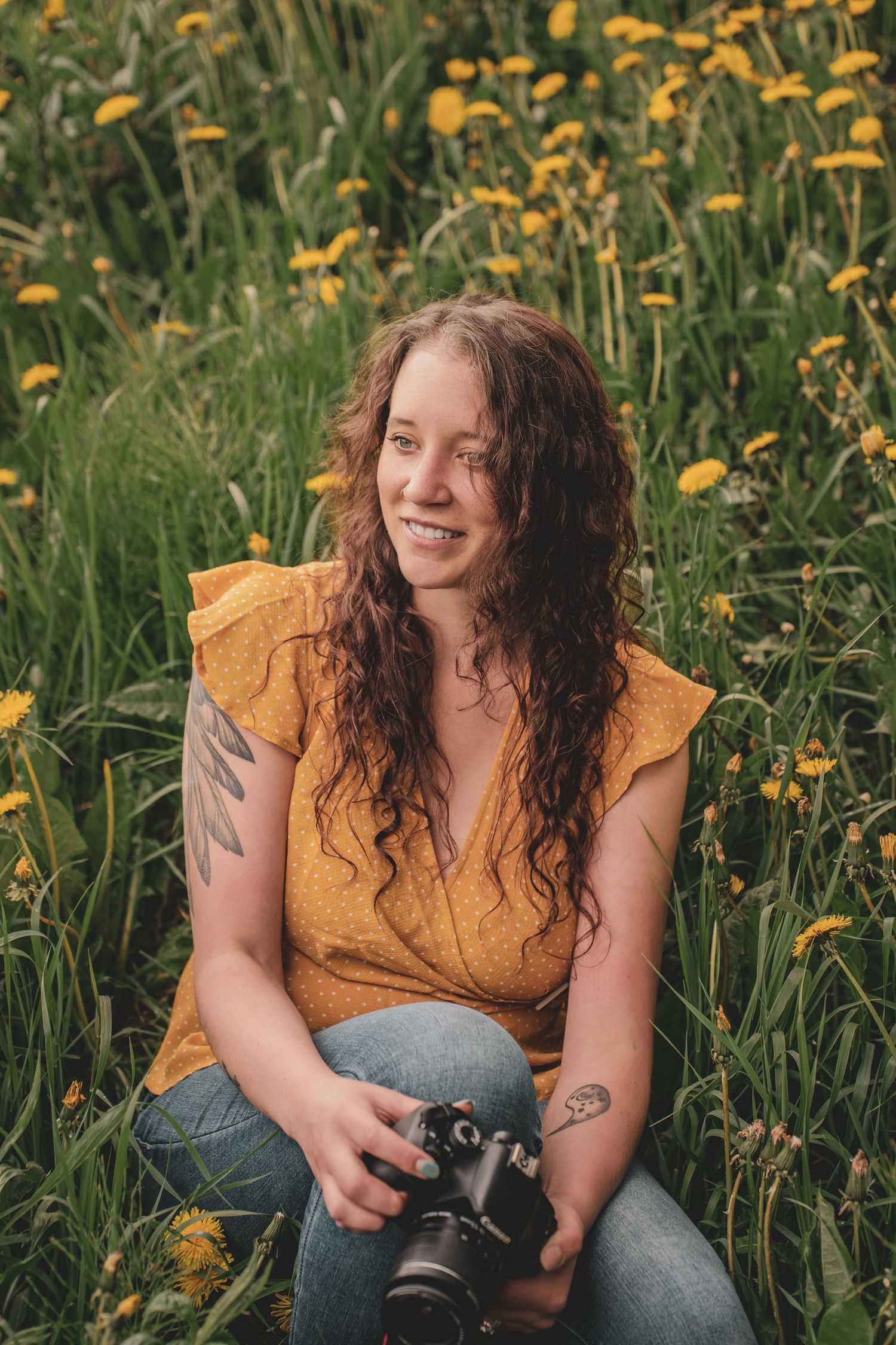 A smiling woman with curly hair holds a camera while sitting in a field of yellow dandelions during photography portfolio photoshoot.