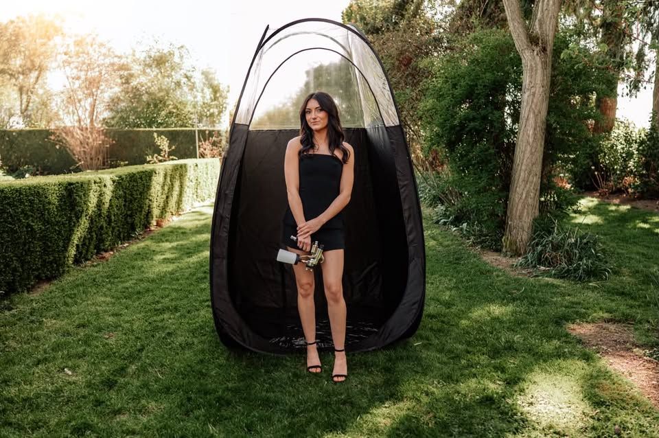 Woman in black mini dress standing beside a black pop-up spray tan tent on a lush green lawn.