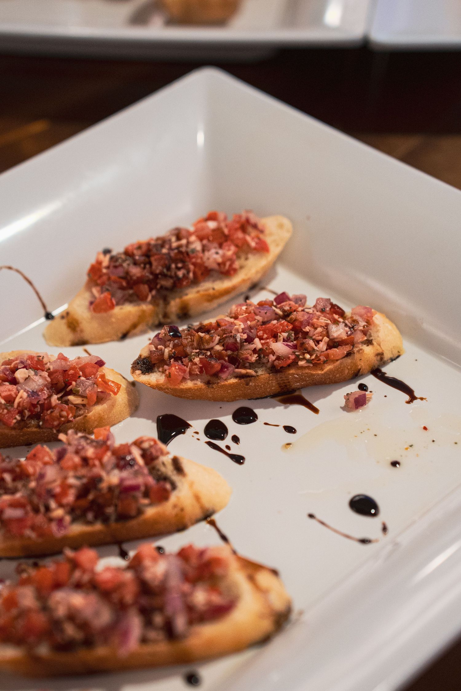 Bruschetta appetizers topped with diced tomatoes and balsamic glaze served on a white rectangular plate.