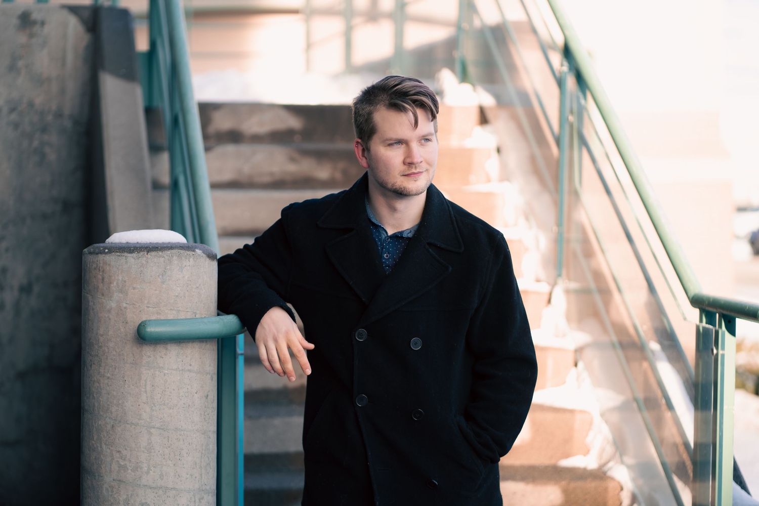 Young actor in black peacoat leaning on railing near outdoor stairs in urban setting during Commercial Business headshots.