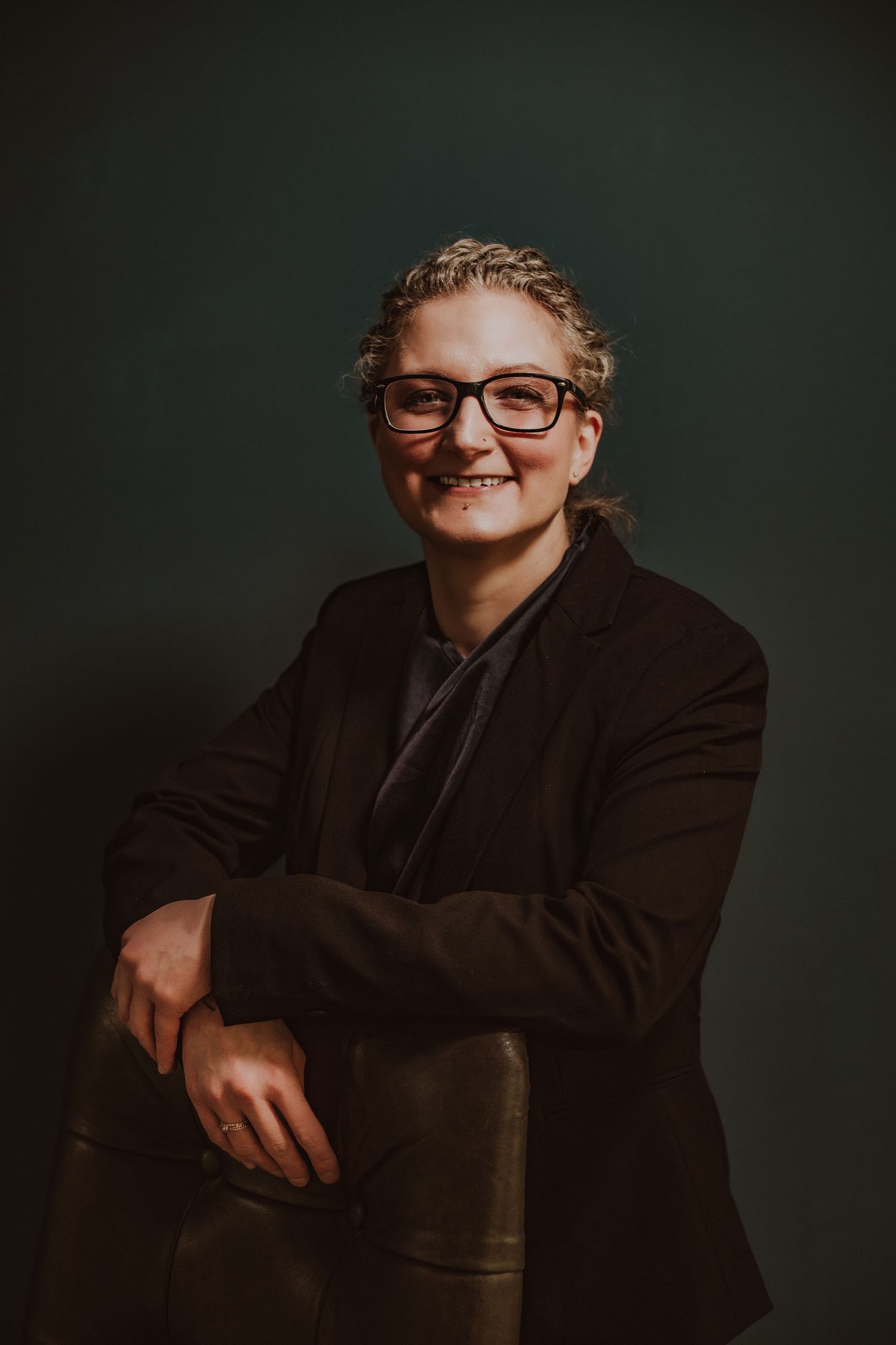 Smiling business woman with braided hair wearing black blazer and red-framed glasses in studio portrait.