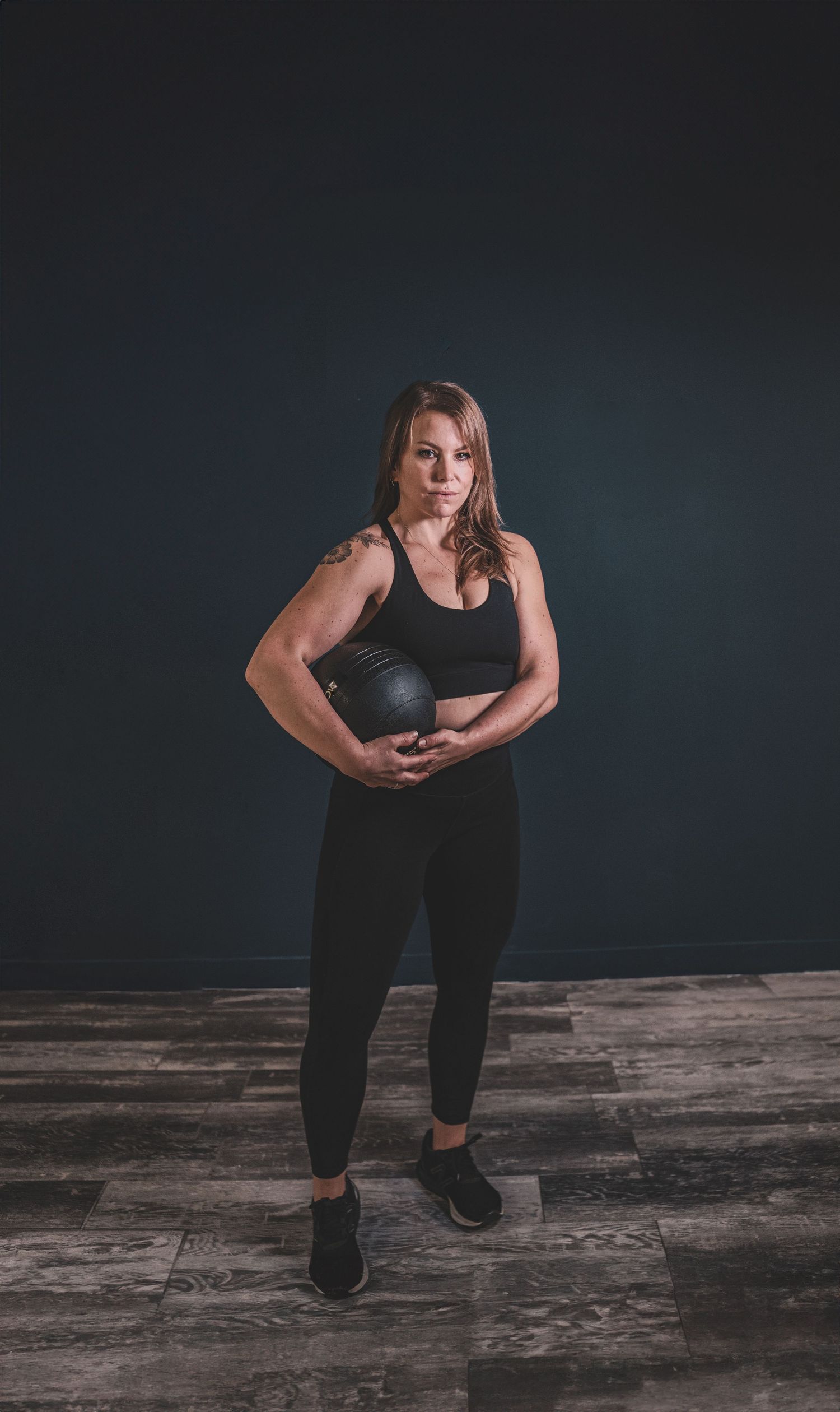A fit woman in black athletic wear holds a medicine ball while standing in a dark fitness studio during a branding photoshoot.