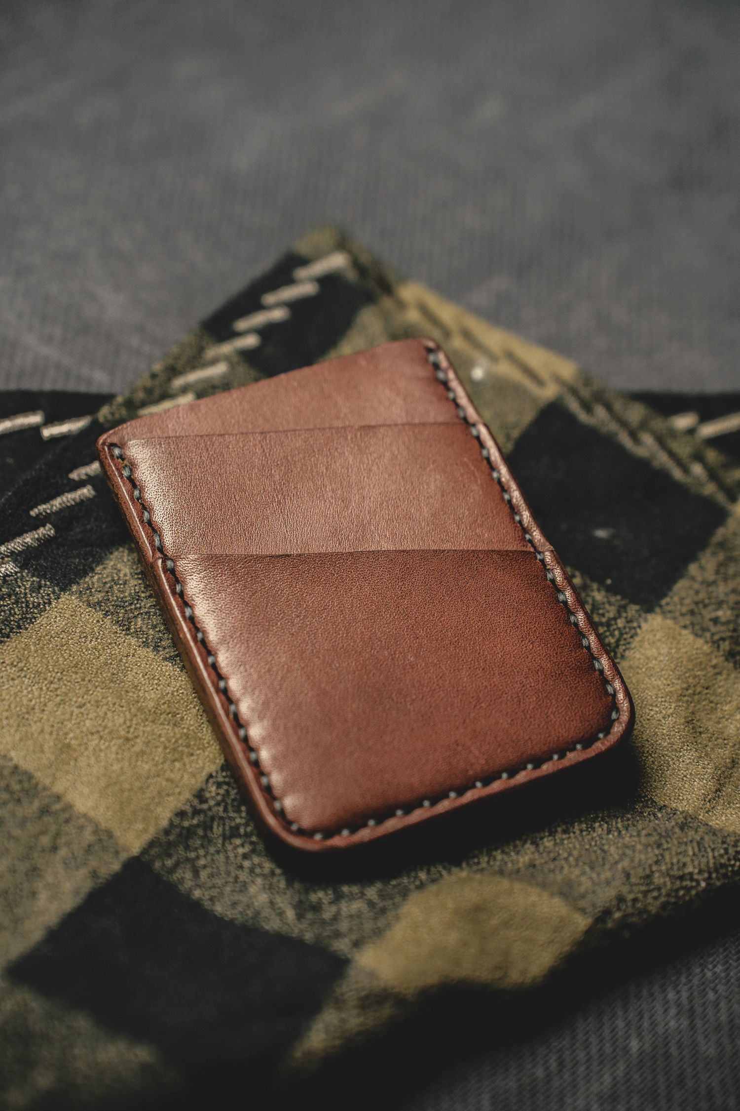 Product Photography of a slim, handstitched brown leather bifold card wallet resting on green camouflage patterned fabric.