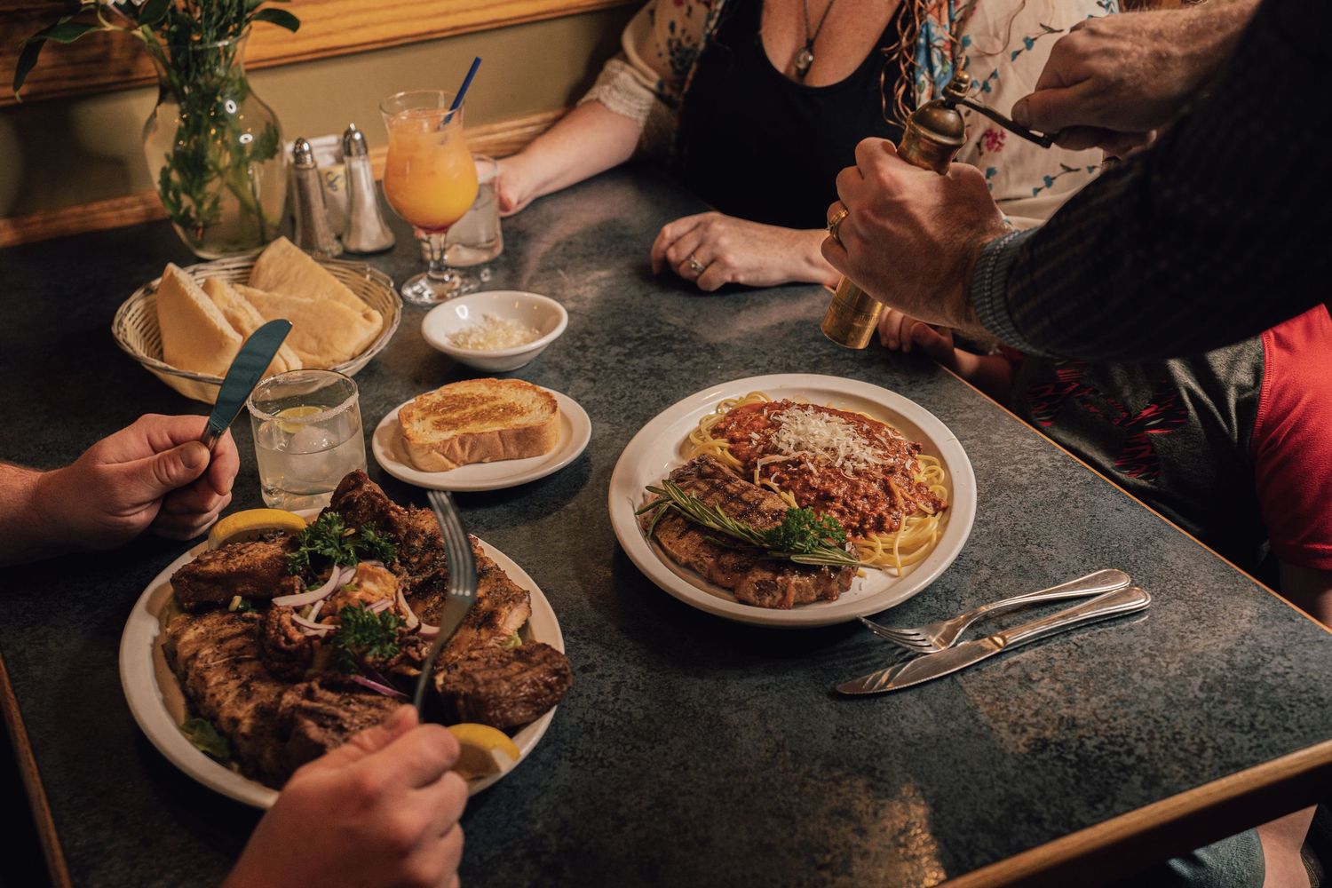 Group of people sharing plates of pasta and grilled meats at a restaurant table.