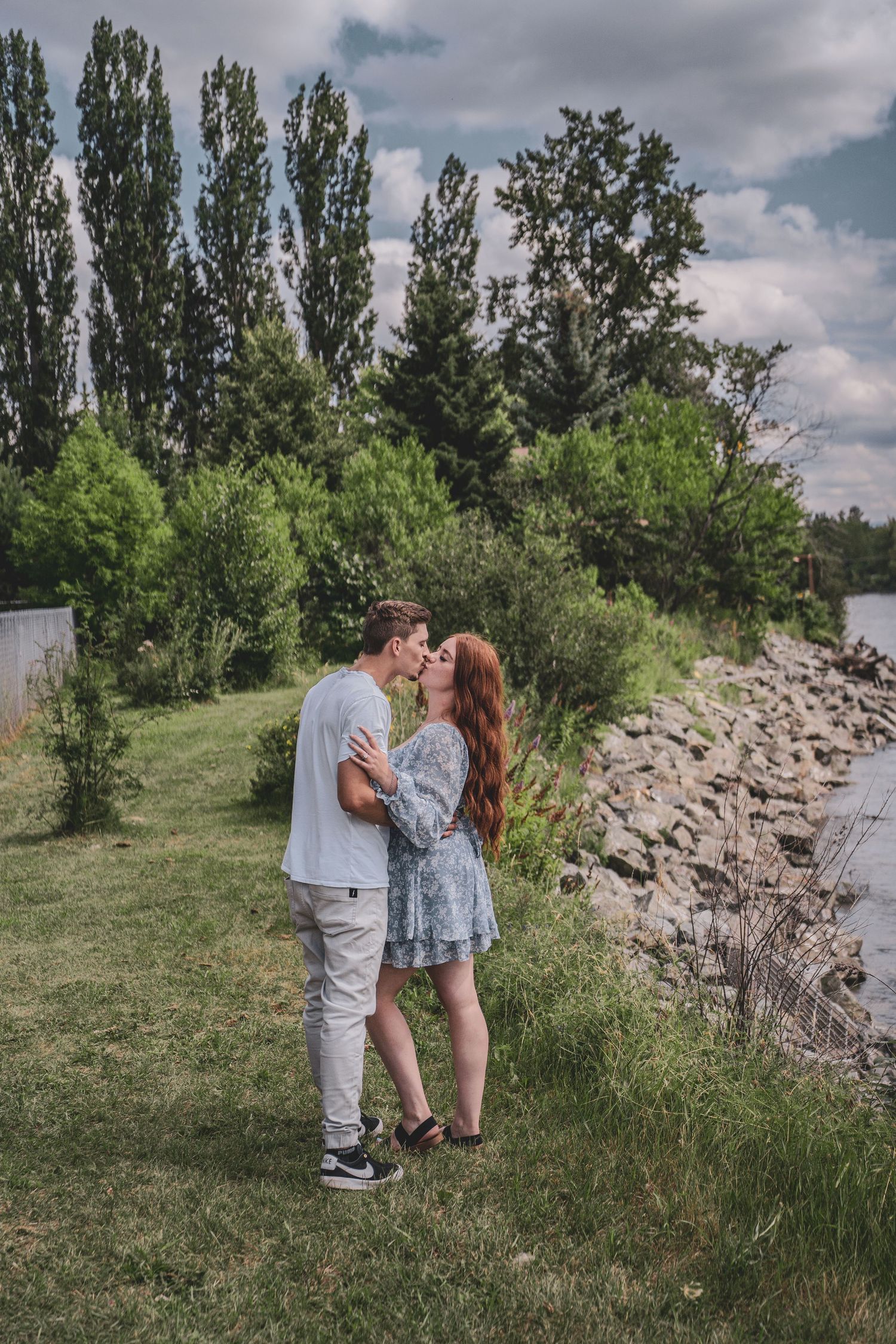 Young couple sharing a kiss on a grassy riverbank surrounded by lush green trees.