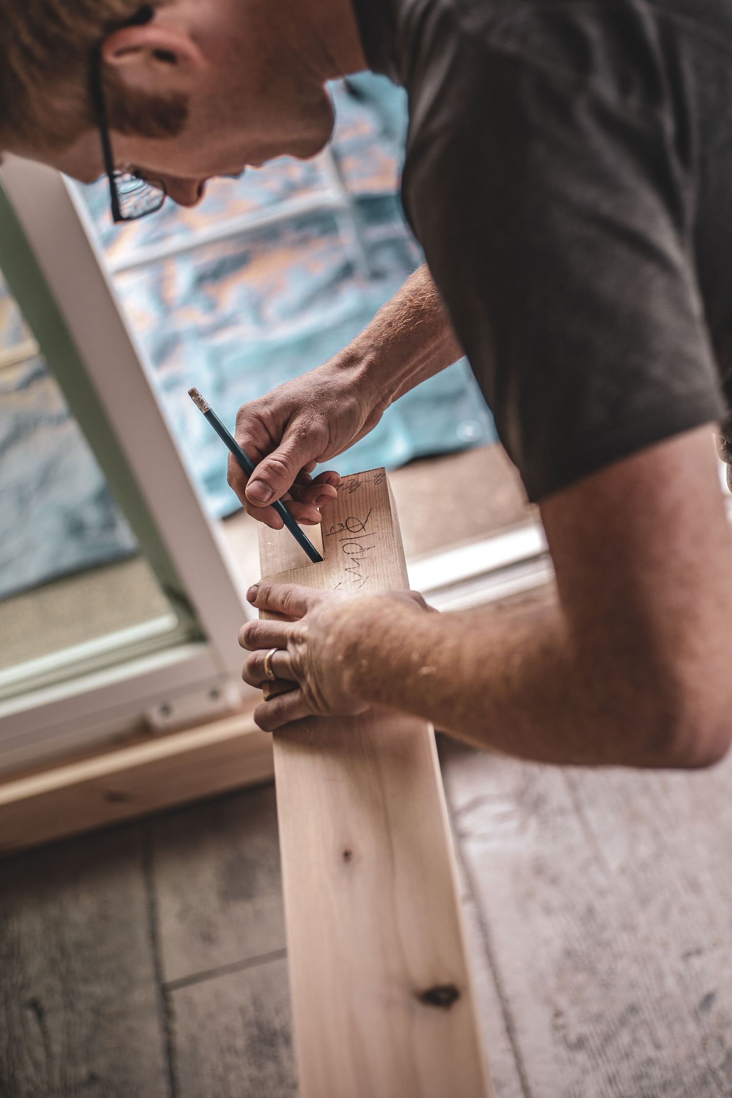 Carpenter marking measurements on wood plank with pencil and square tool near a window during Commercial Business Branding photoshoot.
