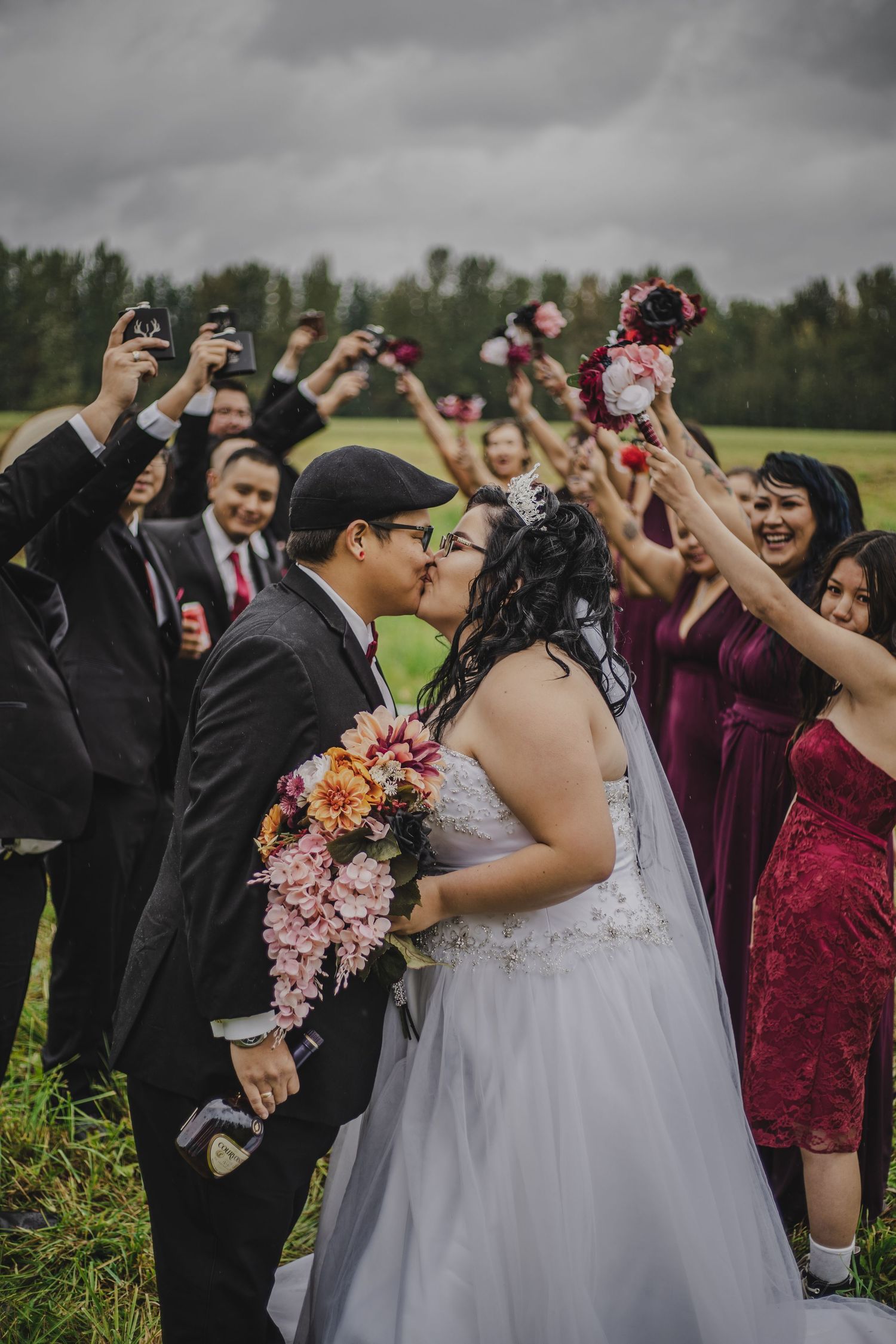 Bride and groom kissing while wedding party cheers with bouquets raised in a green field.