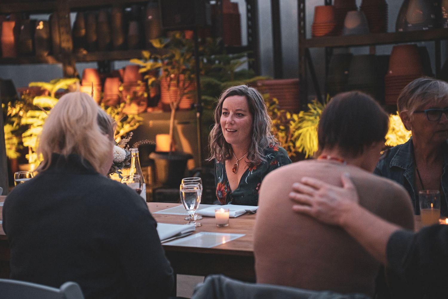 A smiling woman in a floral dress engages in warm conversation at a candlelit dinner table in a greenhouse offered event by tourism pg.