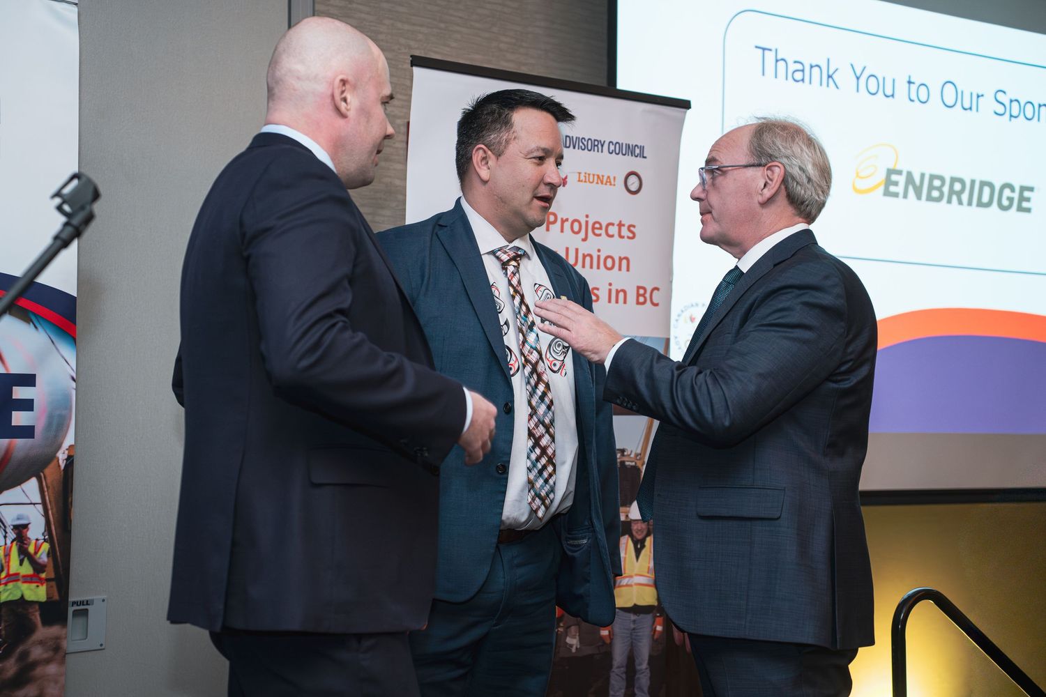 Three businessmen in suits engage in conversation near an Enbridge sponsor banner at a professional conference.