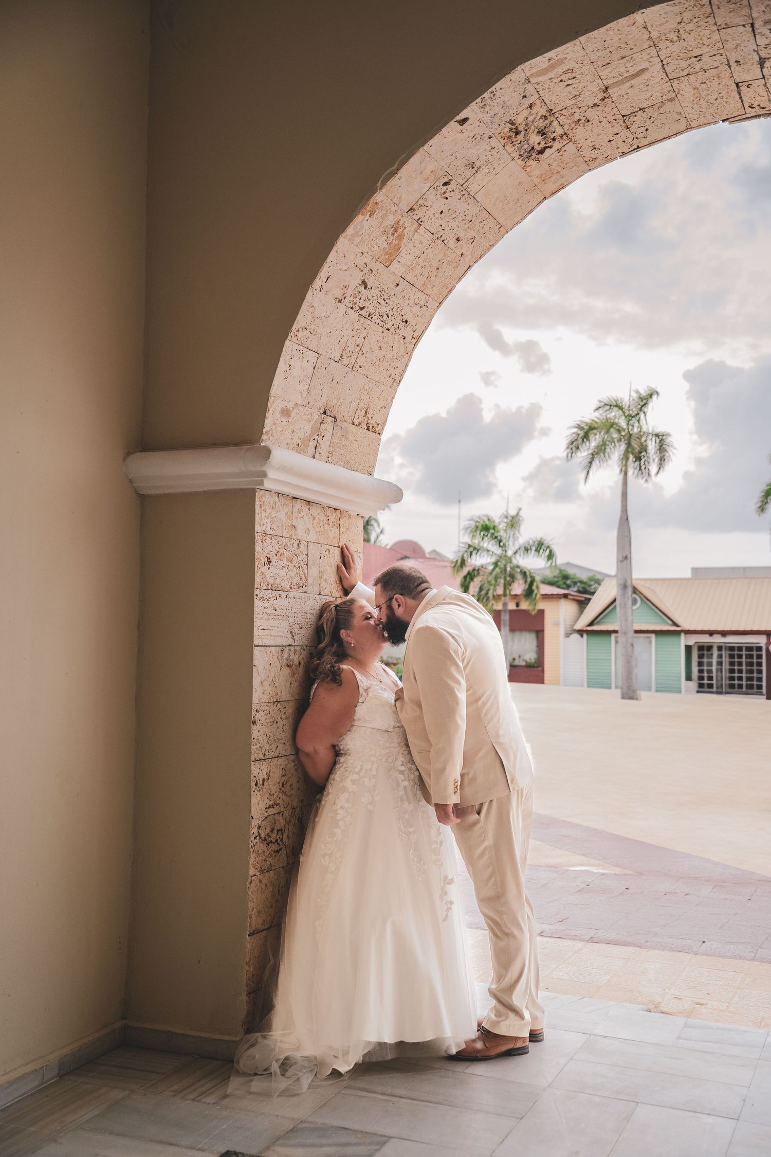 Bride and groom sharing a kiss under a stone arch with palm trees in tropical wedding setting, abroad. Take your photographer abroad!