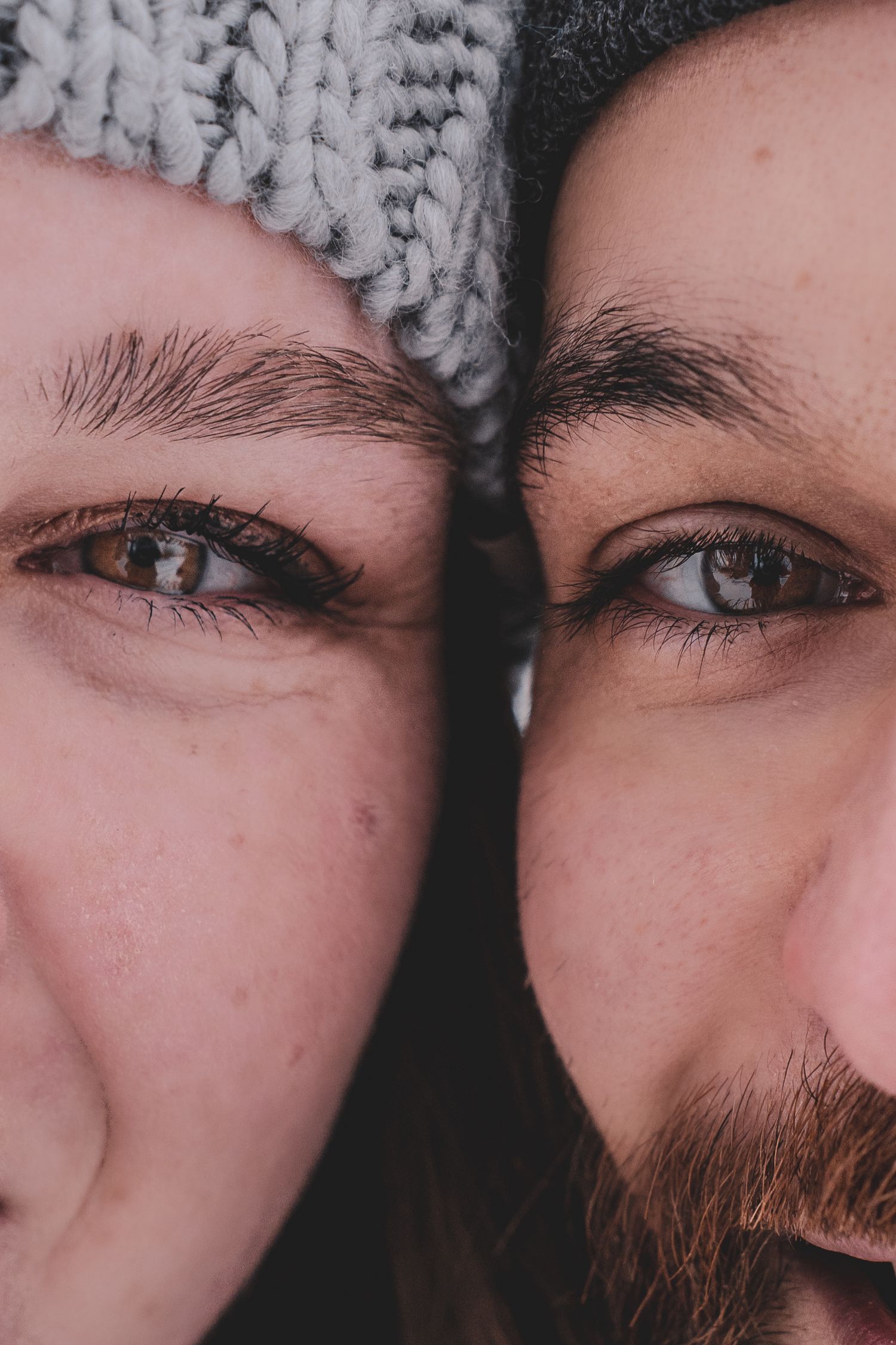 Close-up portrait of couple wearing knit hats with faces together showing brown and blue eyes.