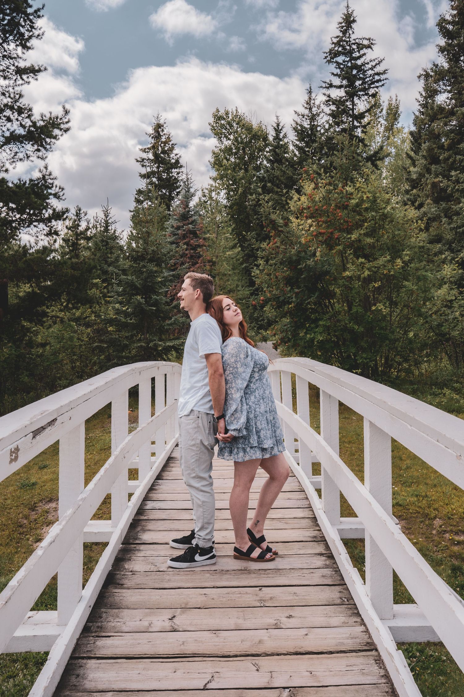 Couple standing back-to-back on a white wooden bridge surrounded by evergreen forest.