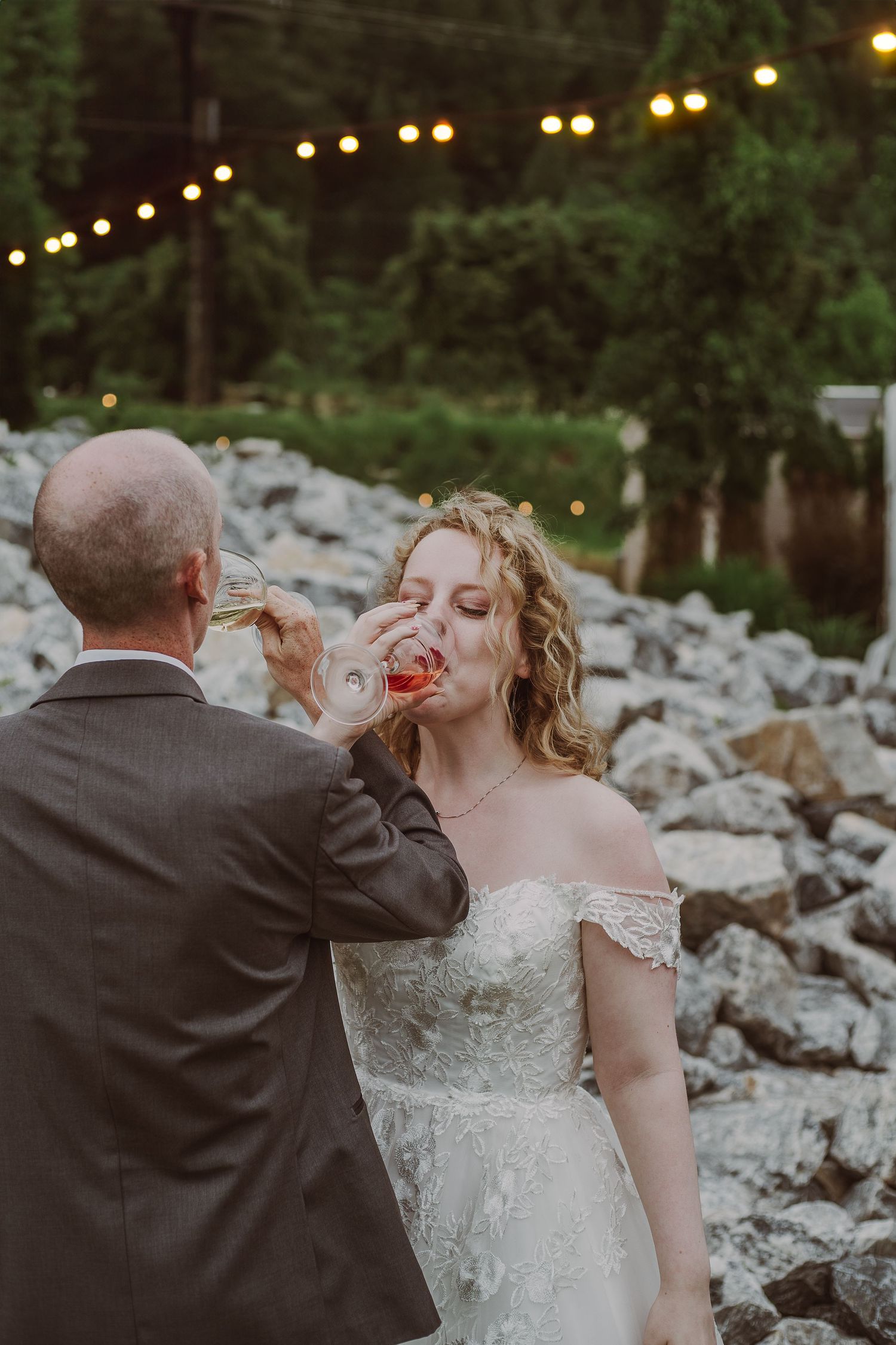 Bride in lace off-shoulder gown drinks from a red shot glass at an outdoor wedding reception with string lights at Northern Lights Winery.