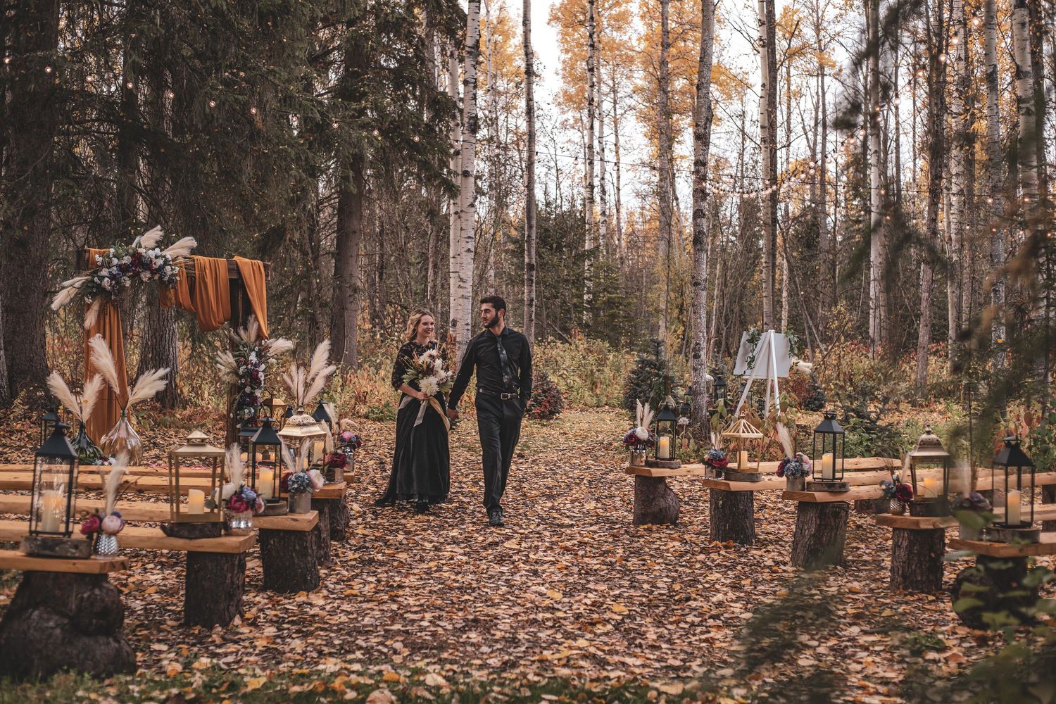 Couple walks hand in hand through a stunning autumn forest wedding ceremony aisle lined with wooden stumps at Willow gardens wedding venue.