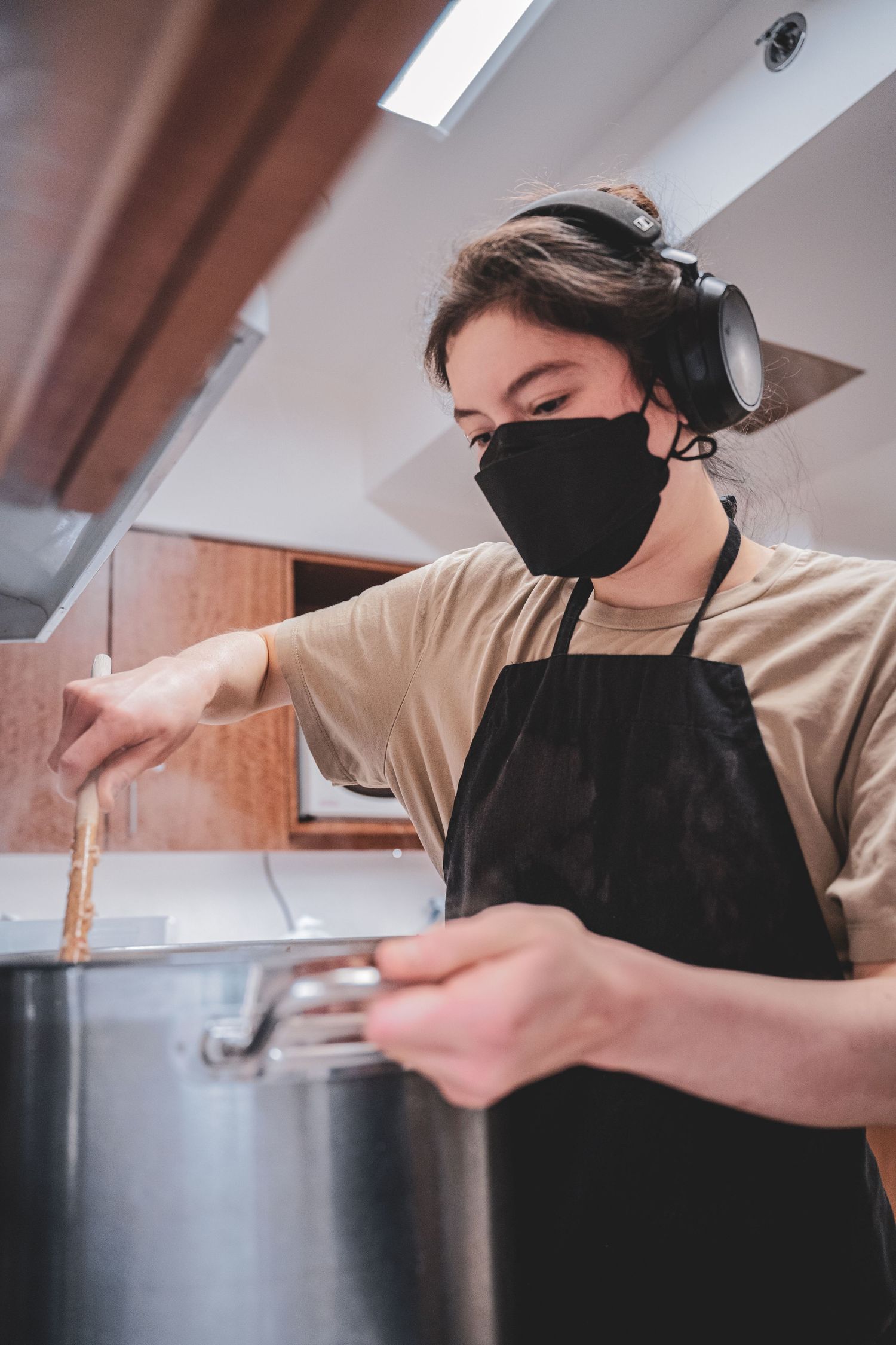 Young person wearing a black face mask, headphones, and apron stirs a large pot in a modern kitchen.