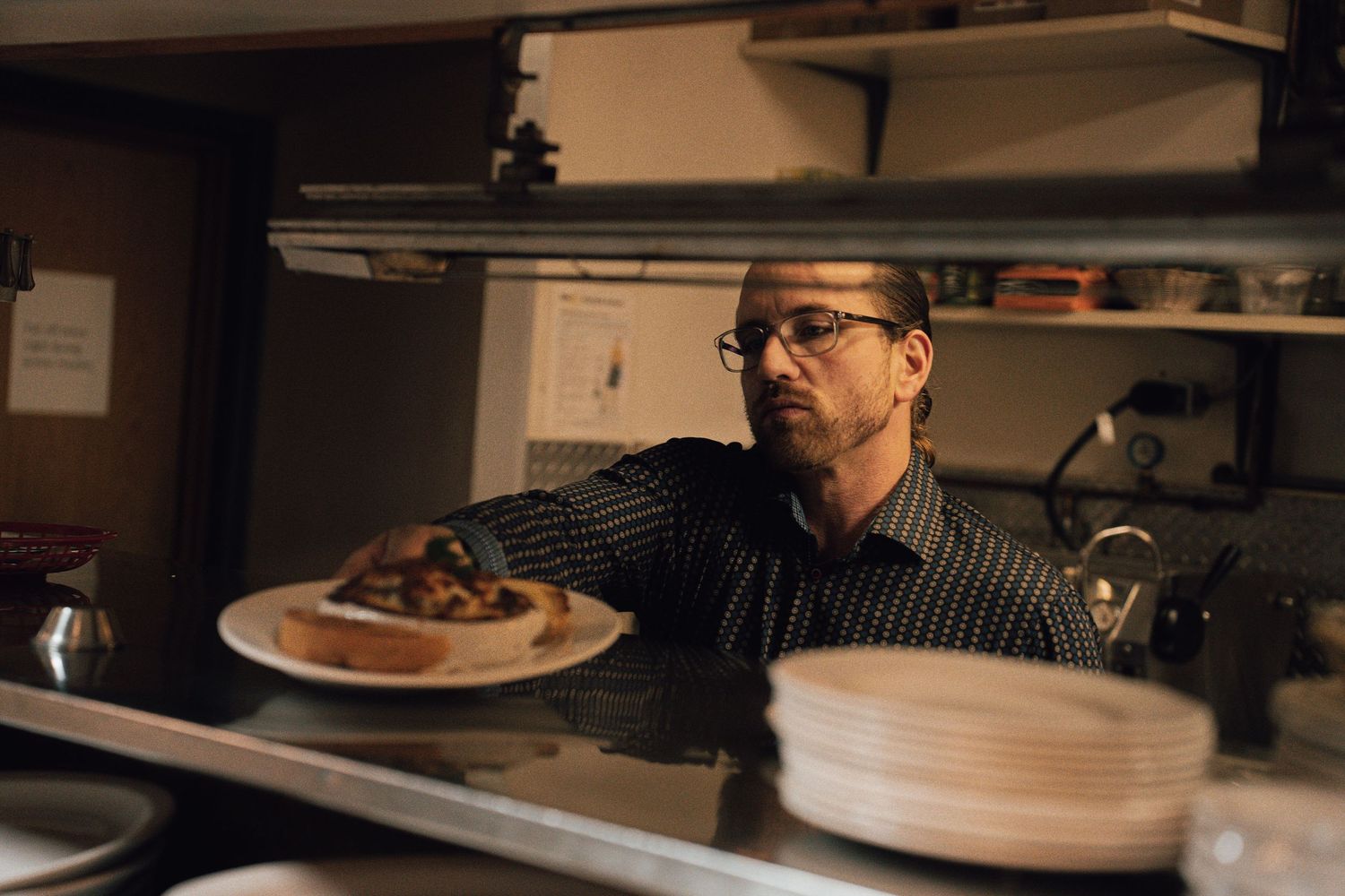Male chef with glasses placing a plated dish on a restaurant pass-through window in a dimly lit kitchen.